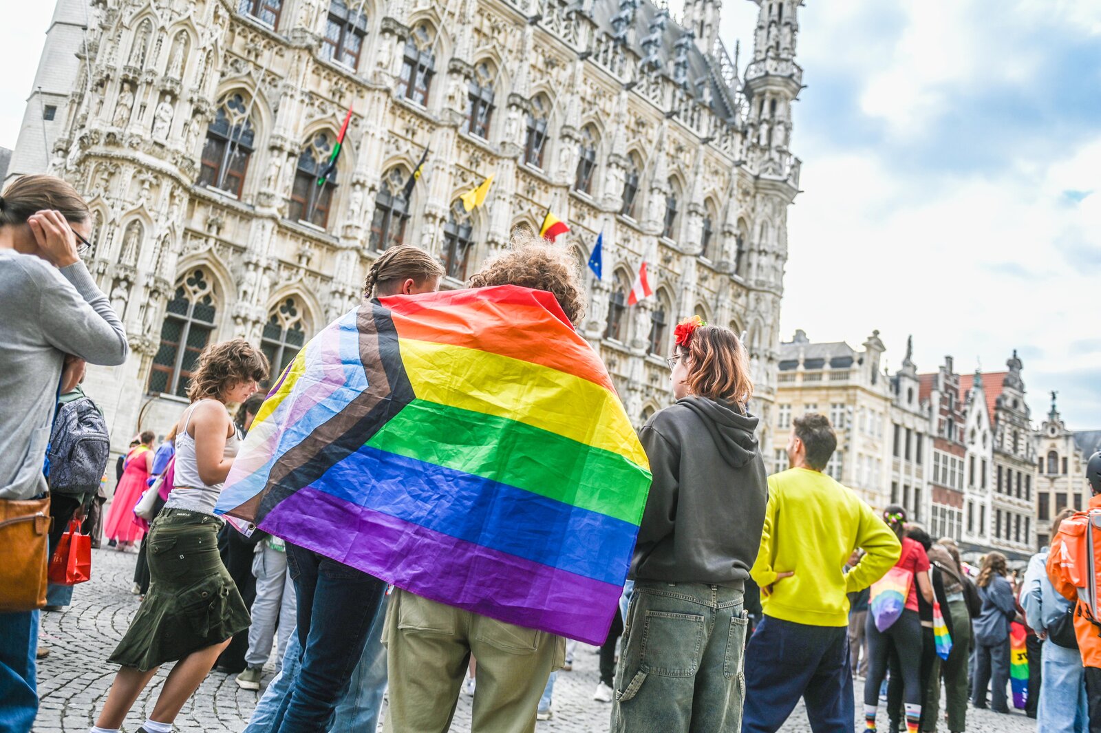 Two people wrapped in one Progress Pride flag in front of the town hall. Leuven Pride 2025, photo: Koen Bauters