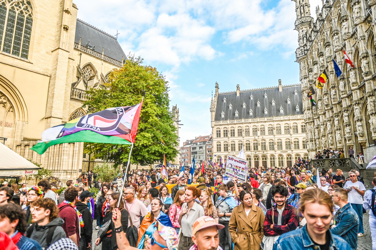 Flags and a placard above the crowd on the square beside the town hall. Leuven Pride 2025, photo: Koen Bauters