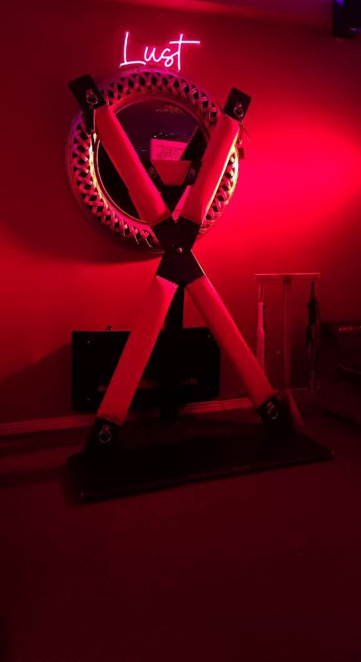 Close-up of a bondage cross (St. Andrew's cross) mounted in front of a round mirror, with a neon "Lust" sign glowing above