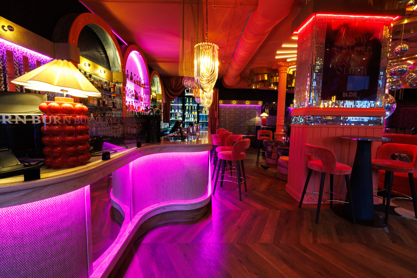 The curved bar counter lit pink from below, with a bubble lamp on the bar, velvet stools and a mirror-tiled pillar