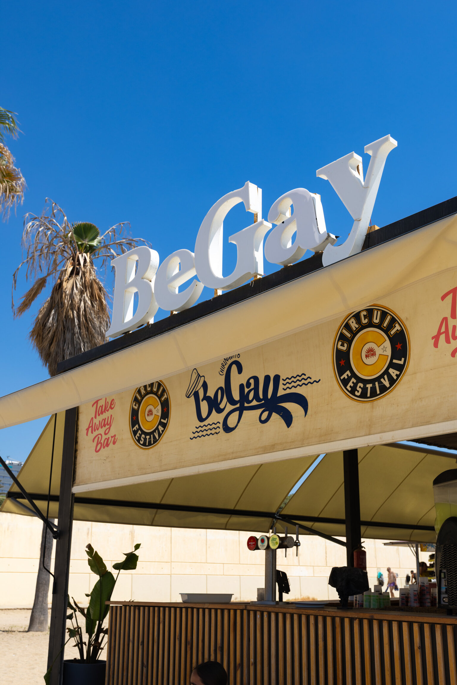 The awning over the bar counter, carrying the BeGay lettering and the Circuit Festival logo, under a palm at the edge of the beach.