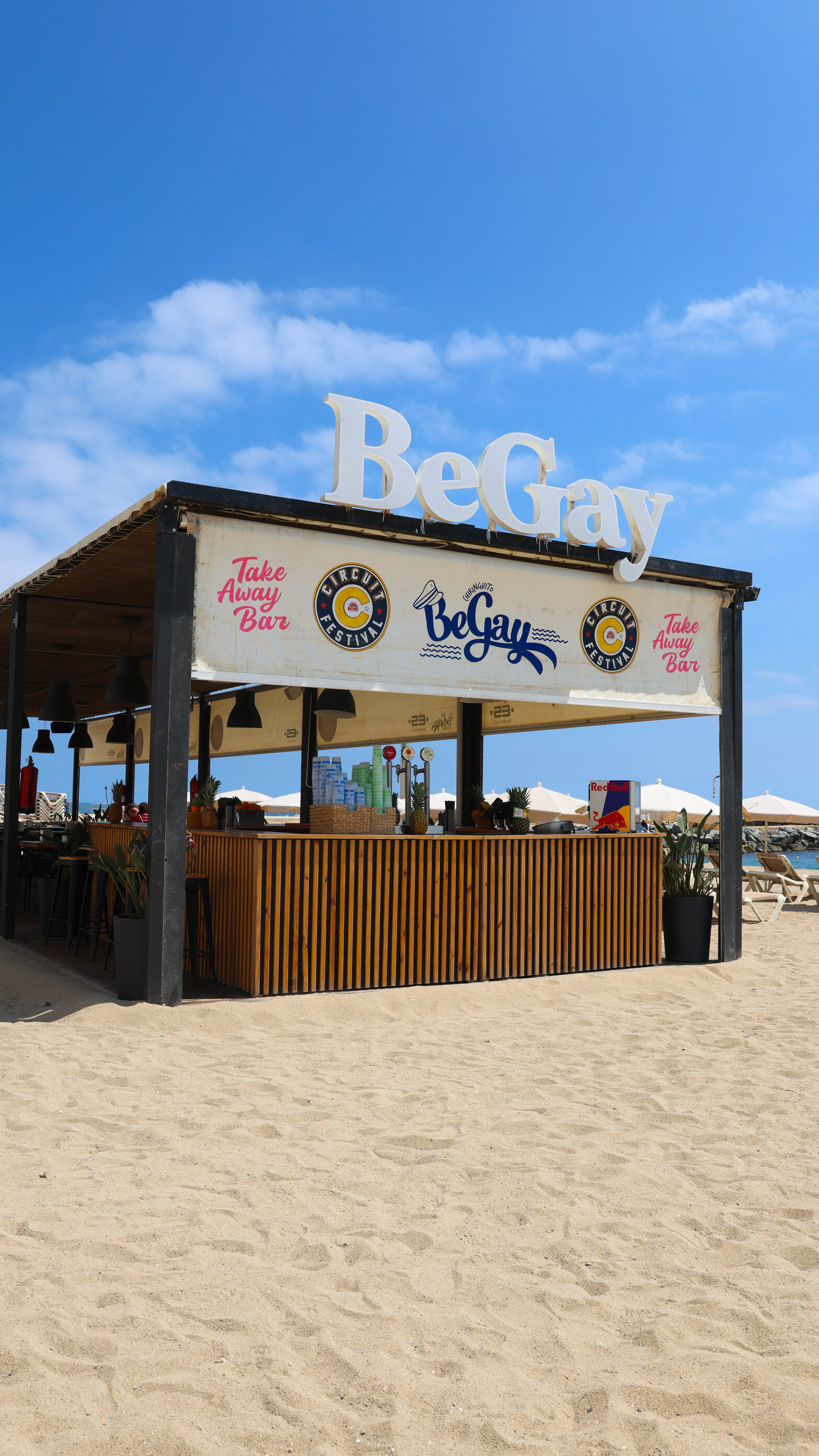 The take away bar standing directly on the sand at La Mar Bella, with the white BeGay lettering above the roof and the loungers and parasols behind it.