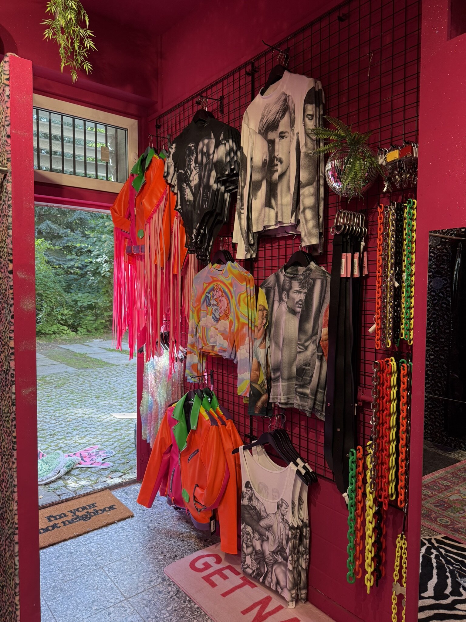 Entrance area of the shop with a pink wall of printed shirts, neon fringed jackets and coloured plastic chains, and the open door onto the courtyard.