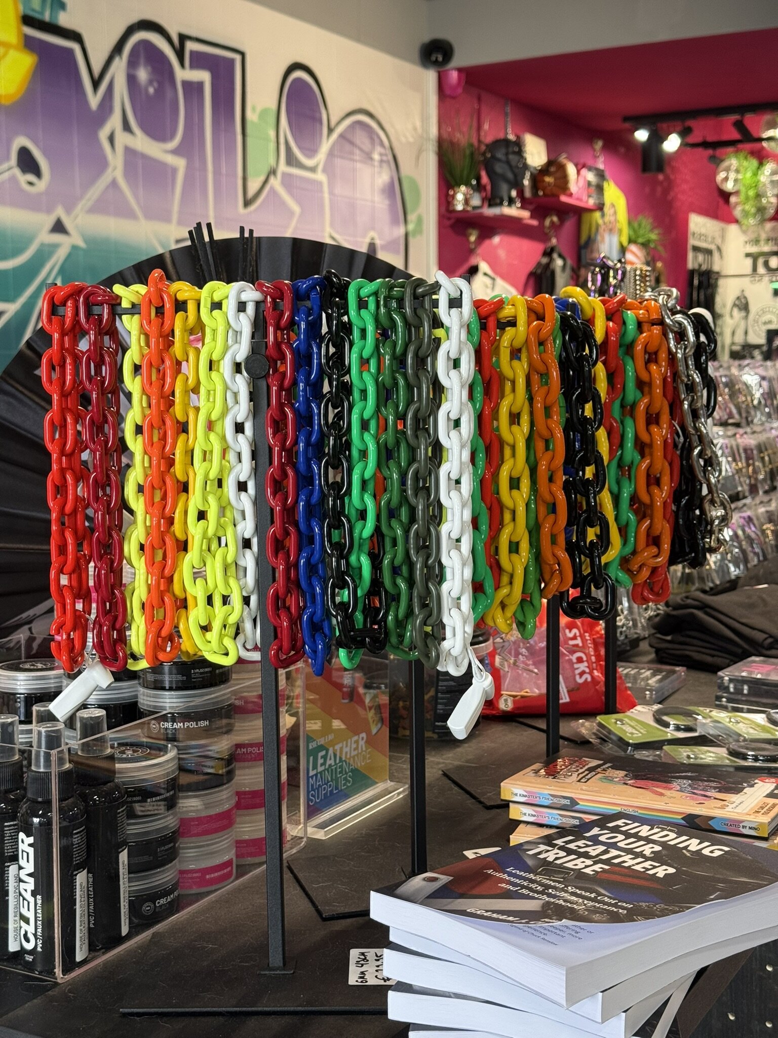 A rack of plastic chains in rainbow, red, black and green next to leather care products and the book Finding Your Leather Tribe, with the graffiti mural behind.