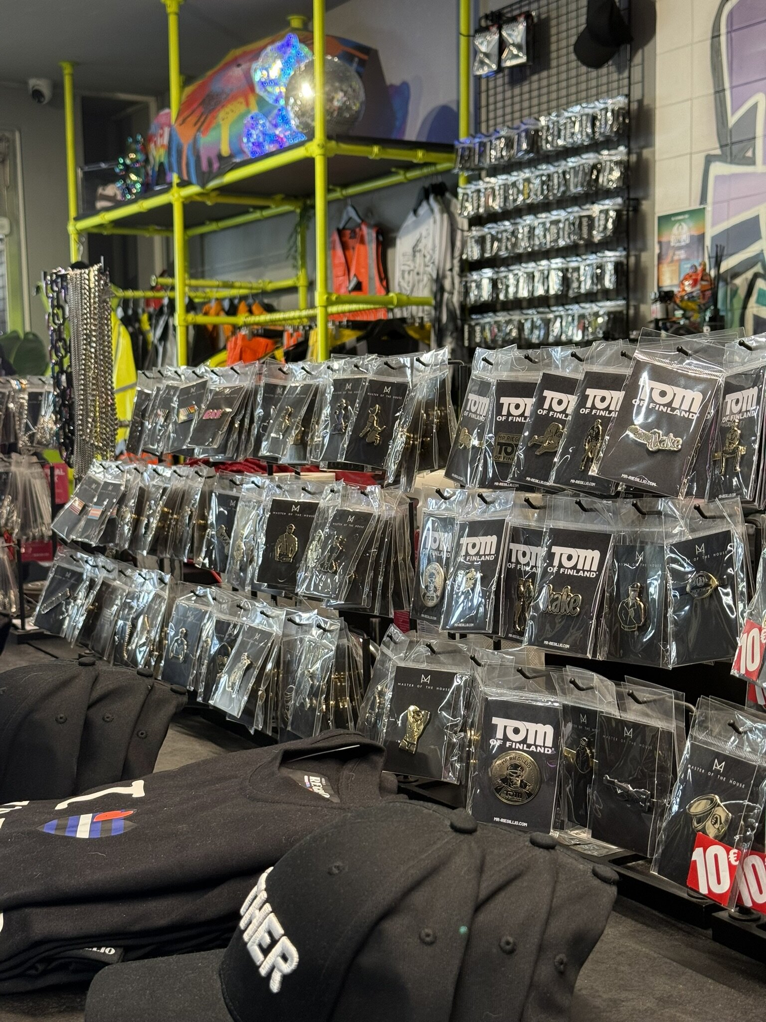 Display of enamel pins and badges, including Tom of Finland designs, in front of a lime green shelving frame.