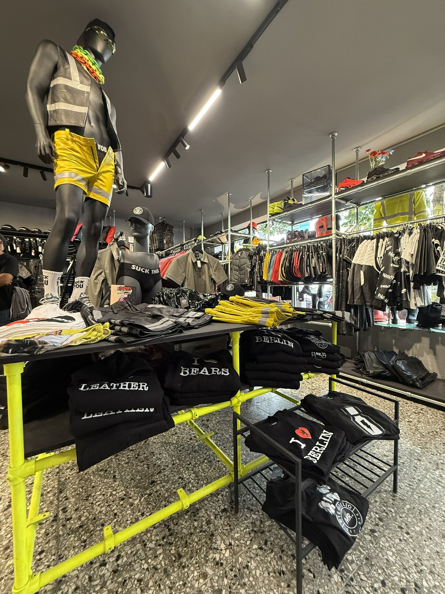 Main room with a mannequin in a reflective vest and yellow shorts, folded Berlin and leather T-shirts on a table, and jackets and shorts on rails along the wall.