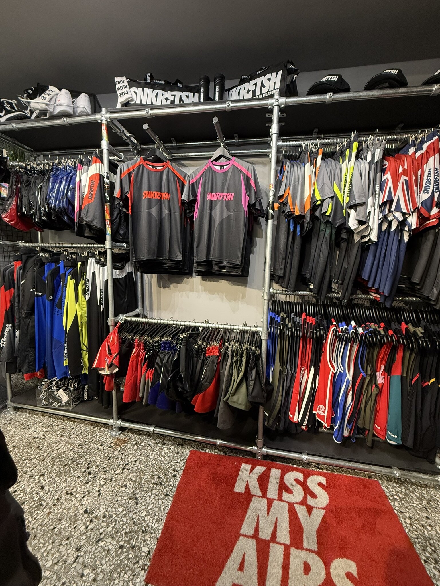 Wall of sportswear on steel rails, with jerseys, shorts and caps from the SNKRFTSH label above a red doormat.