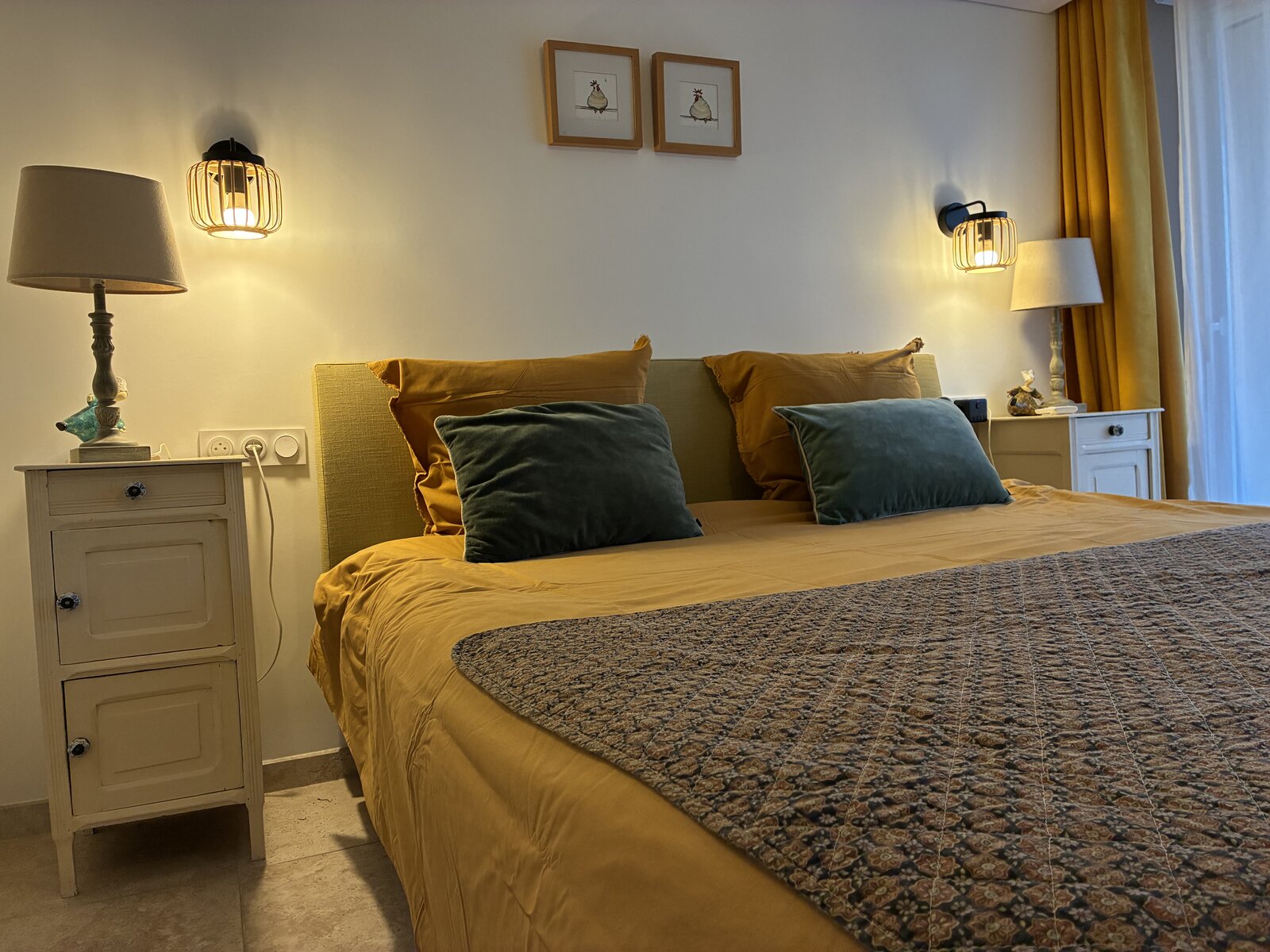 A bedroom in mustard and sage tones, with wall lamps either side of the bed and two rooster prints above the headboard.