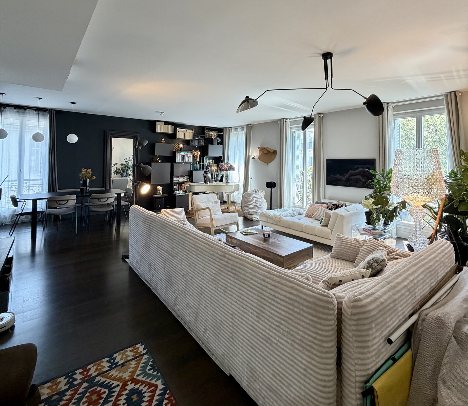 Open-plan living room with a long striped corduroy sofa, a wooden coffee table, a dining table by the windows and a dark shelf wall at the back.