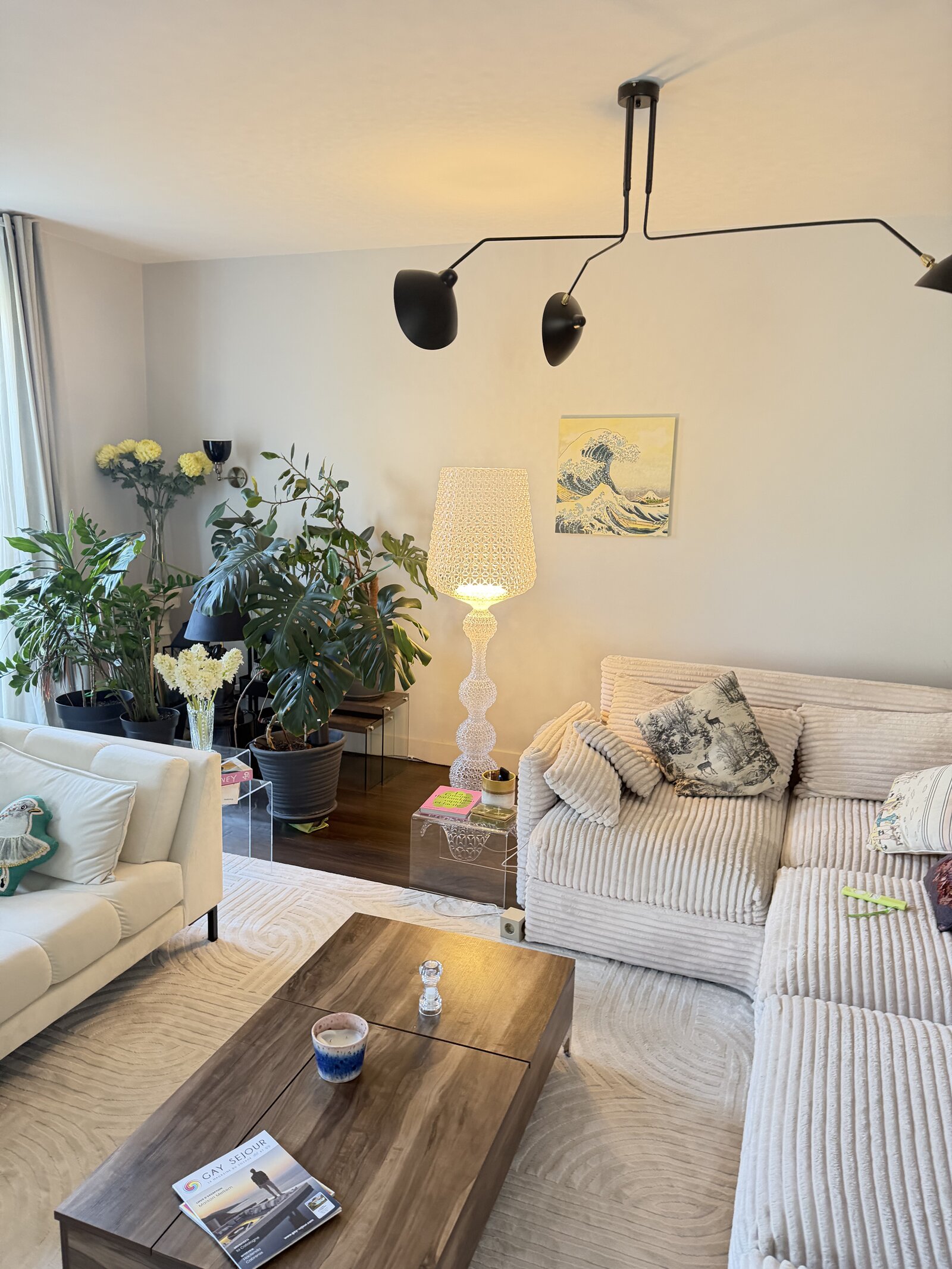 Living room corner with a white corduroy sofa, a wooden coffee table, large potted plants and a tall white lace-patterned floor lamp.