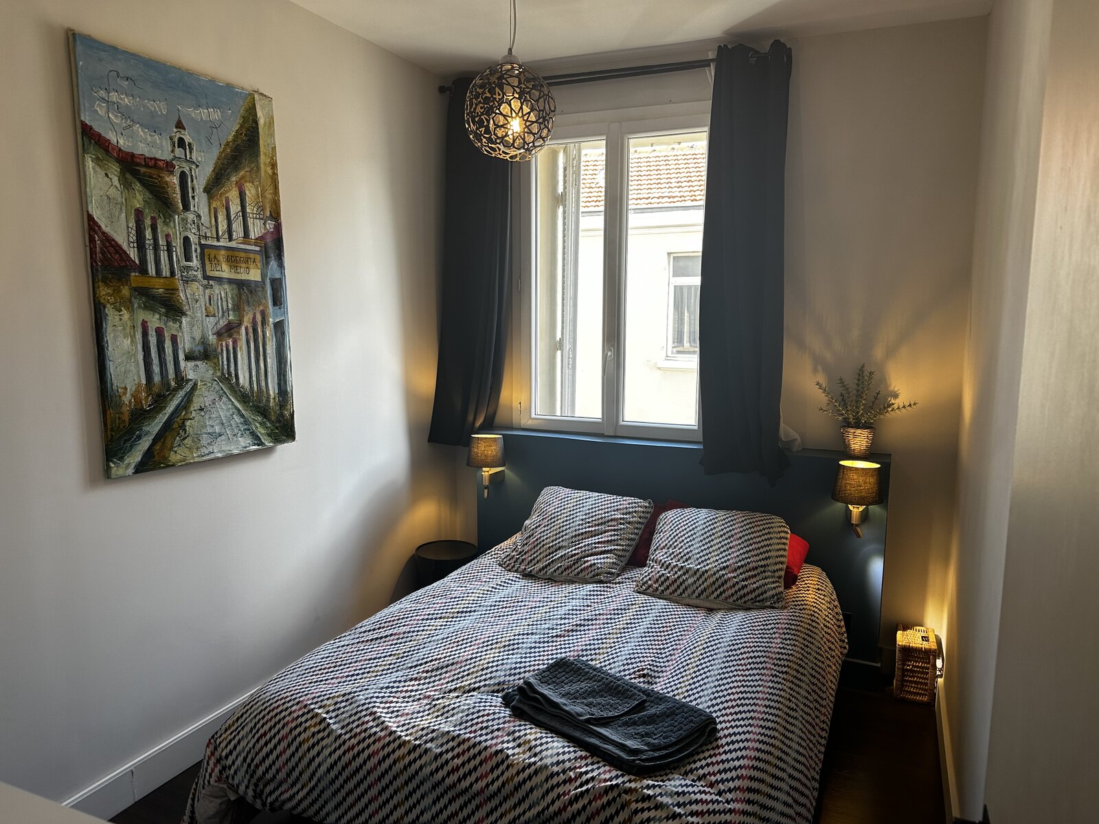 Double bedroom with a patterned bedspread, two bedside lamps, dark curtains at the window and a painting of an old street on the wall.