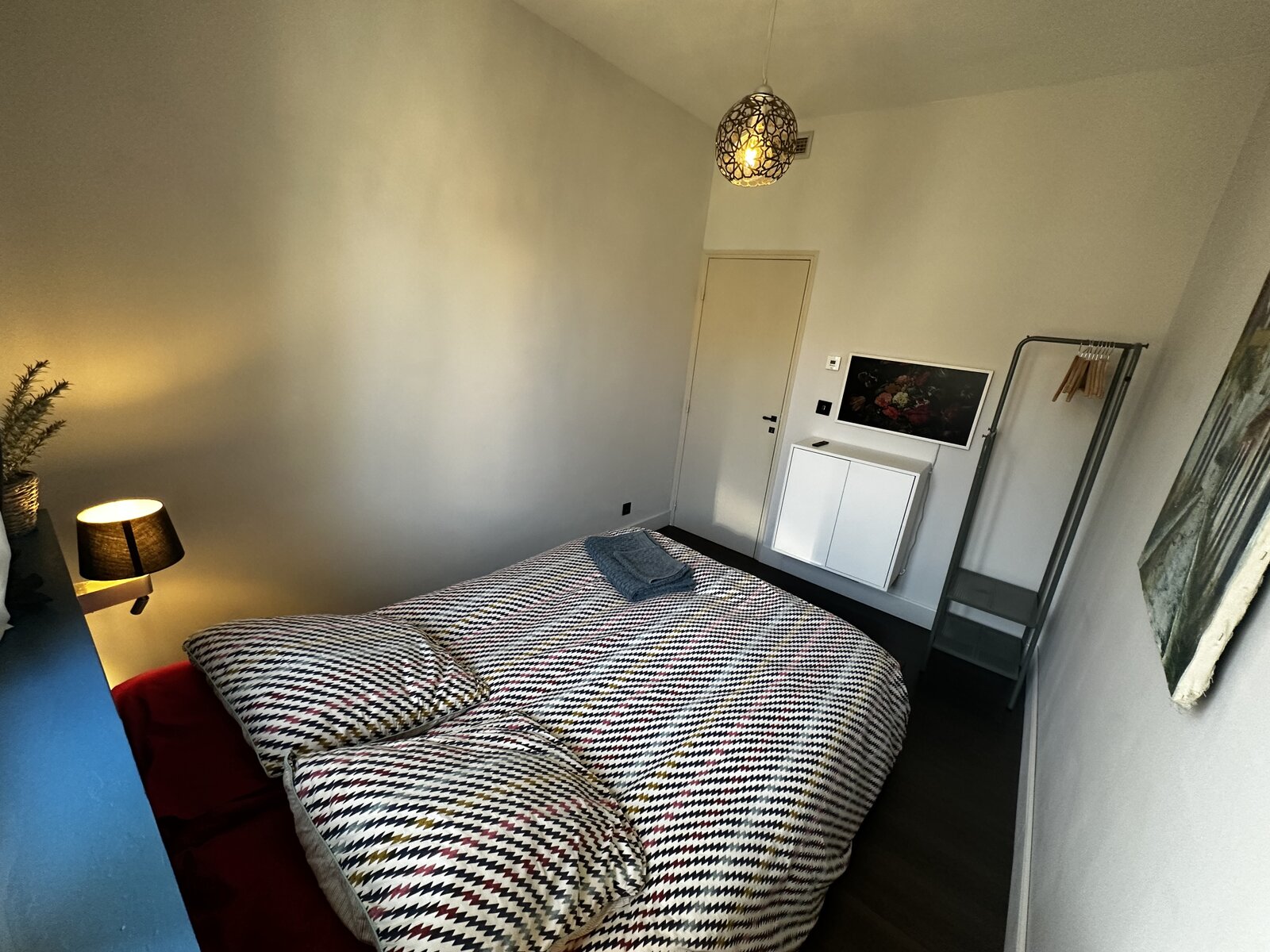 Double bedroom with a patterned bedspread and folded towels, a pendant lamp, a small white cabinet and a clothes rail by the door.