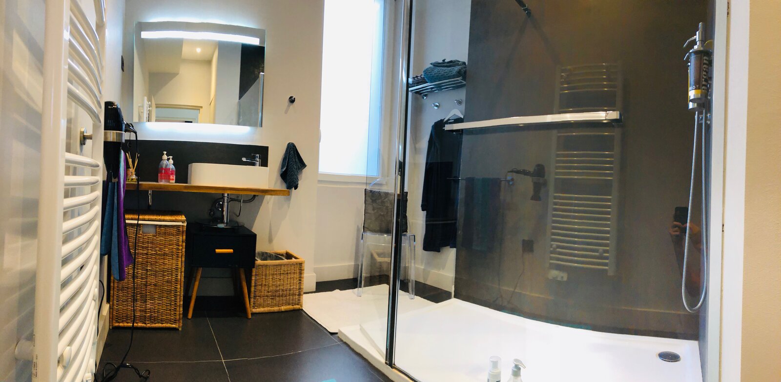 Bathroom with a walk-in glass shower, a towel radiator, a vessel basin under a lit mirror and wicker baskets.