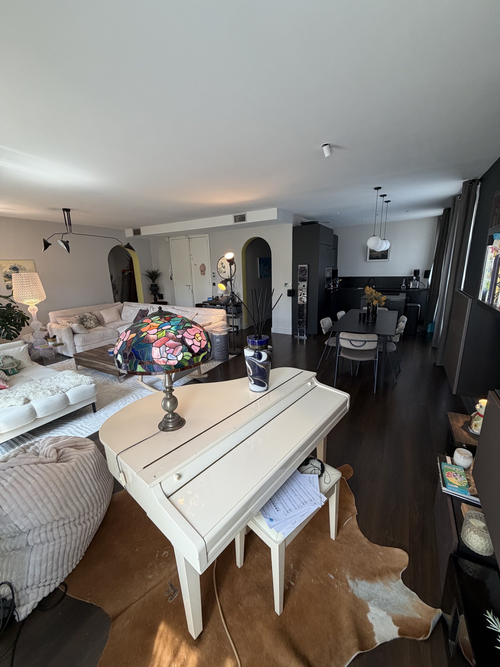 Long living and dining space with a white baby grand piano and a stained-glass lamp in front, sofas on the left and a dining table on the right.