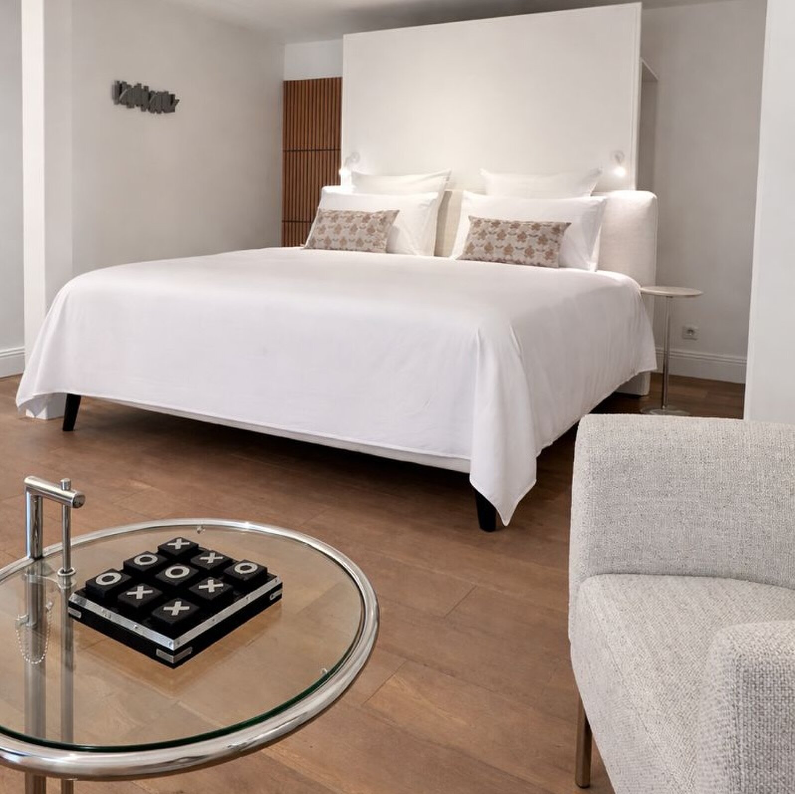 Bedroom with a large white bed, a round glass side table with a noughts and crosses game and a grey armchair in the foreground.