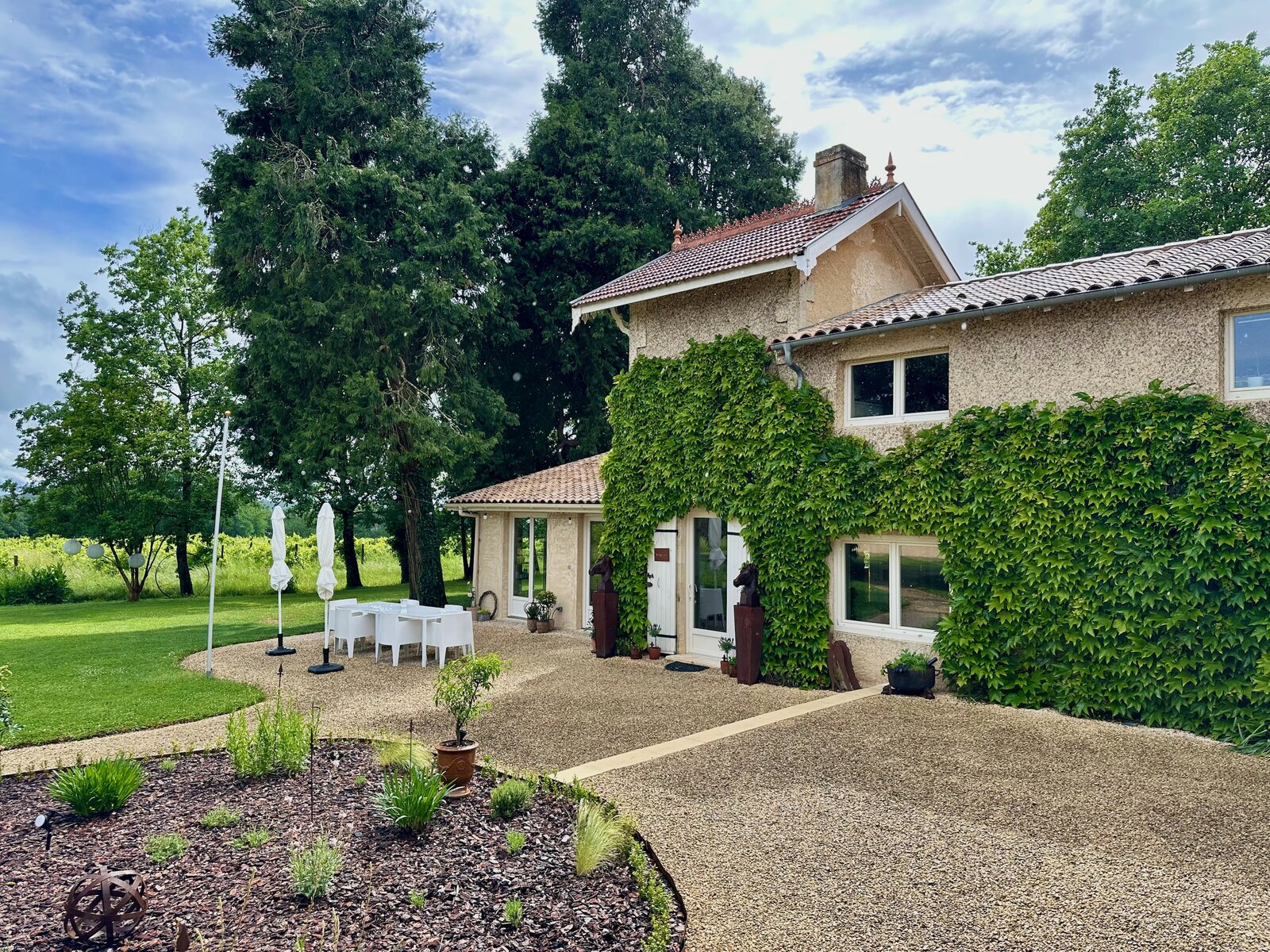 The ivy-covered house with its gravel courtyard, a planted bed in front and a terrace with white chairs and parasols by the lawn.