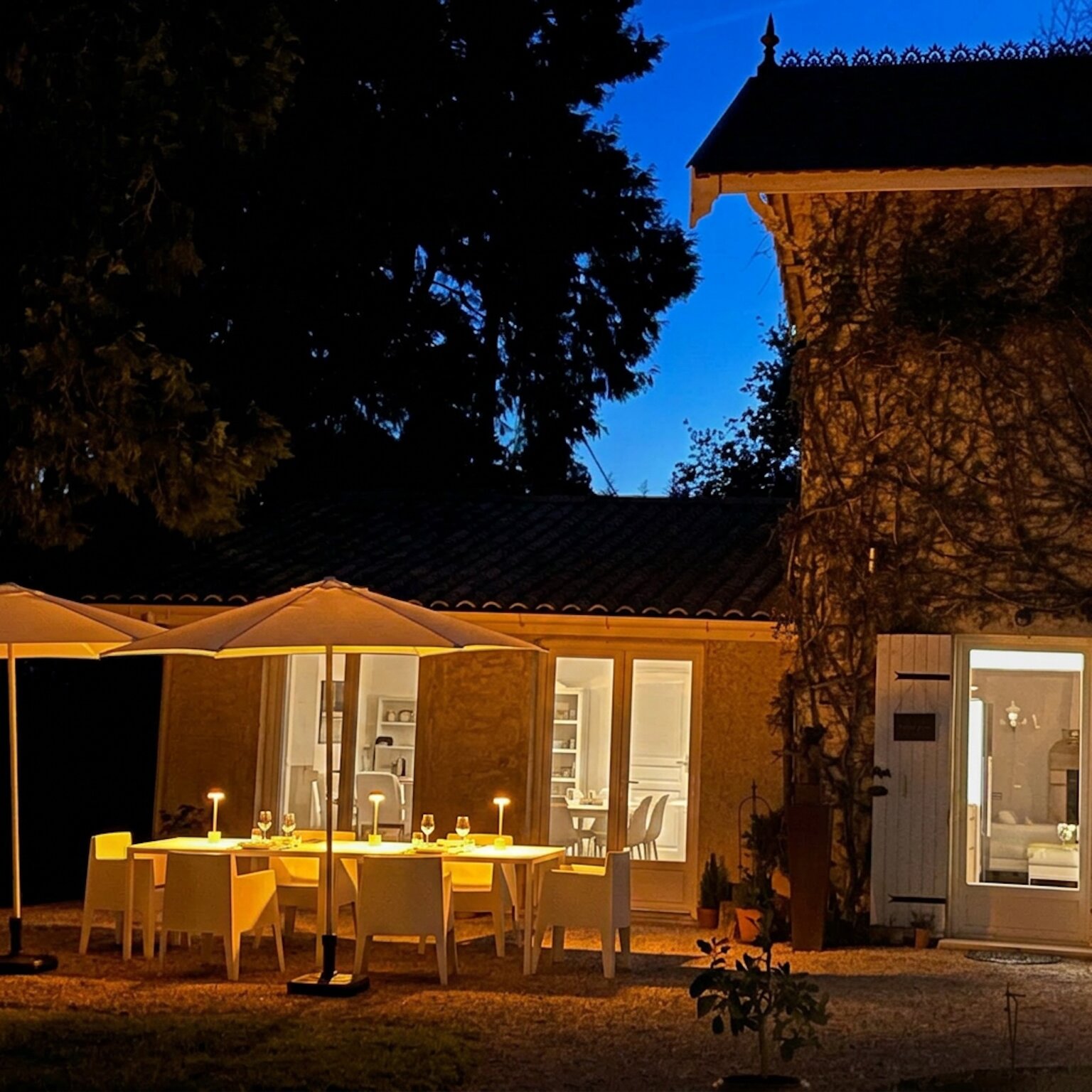 The terrace at night, a table with small lamps under two parasols and the lit windows of the house behind.