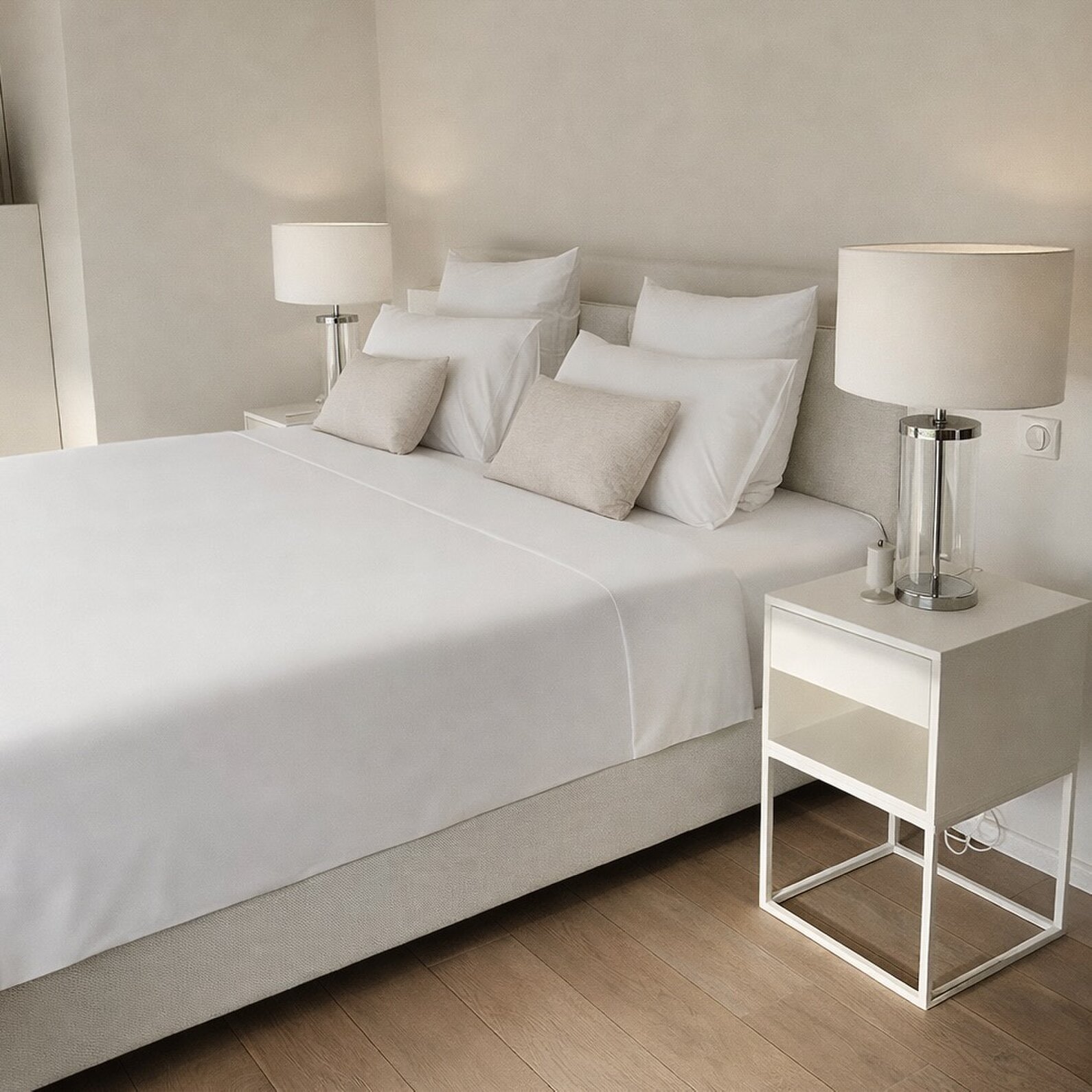 Double bed with white linen and beige cushions, flanked by two glass table lamps on white bedside tables.