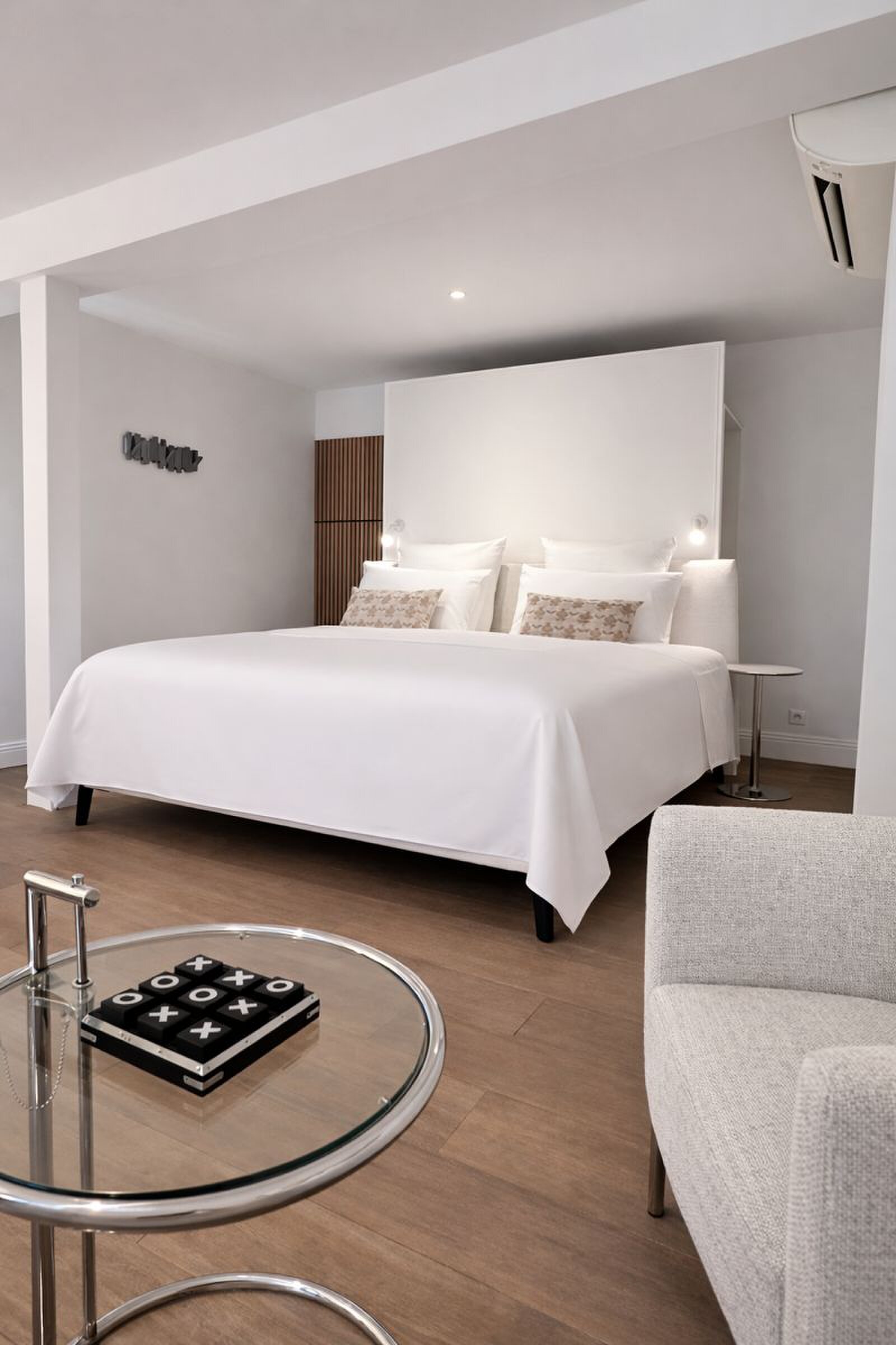 Bedroom with a large white bed against a freestanding headboard wall, a glass side table with a noughts and crosses game and a grey armchair.