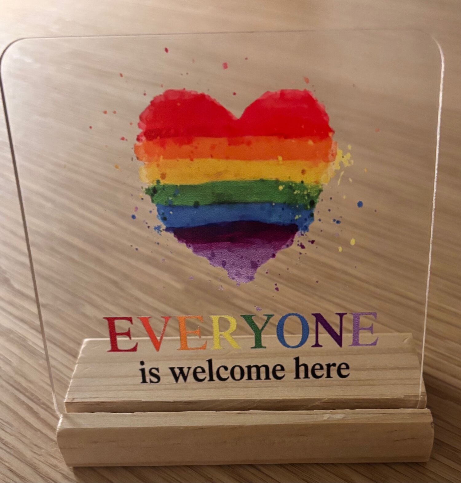 A small acrylic sign with a rainbow heart and the words "Everyone is welcome here" on a wooden stand.