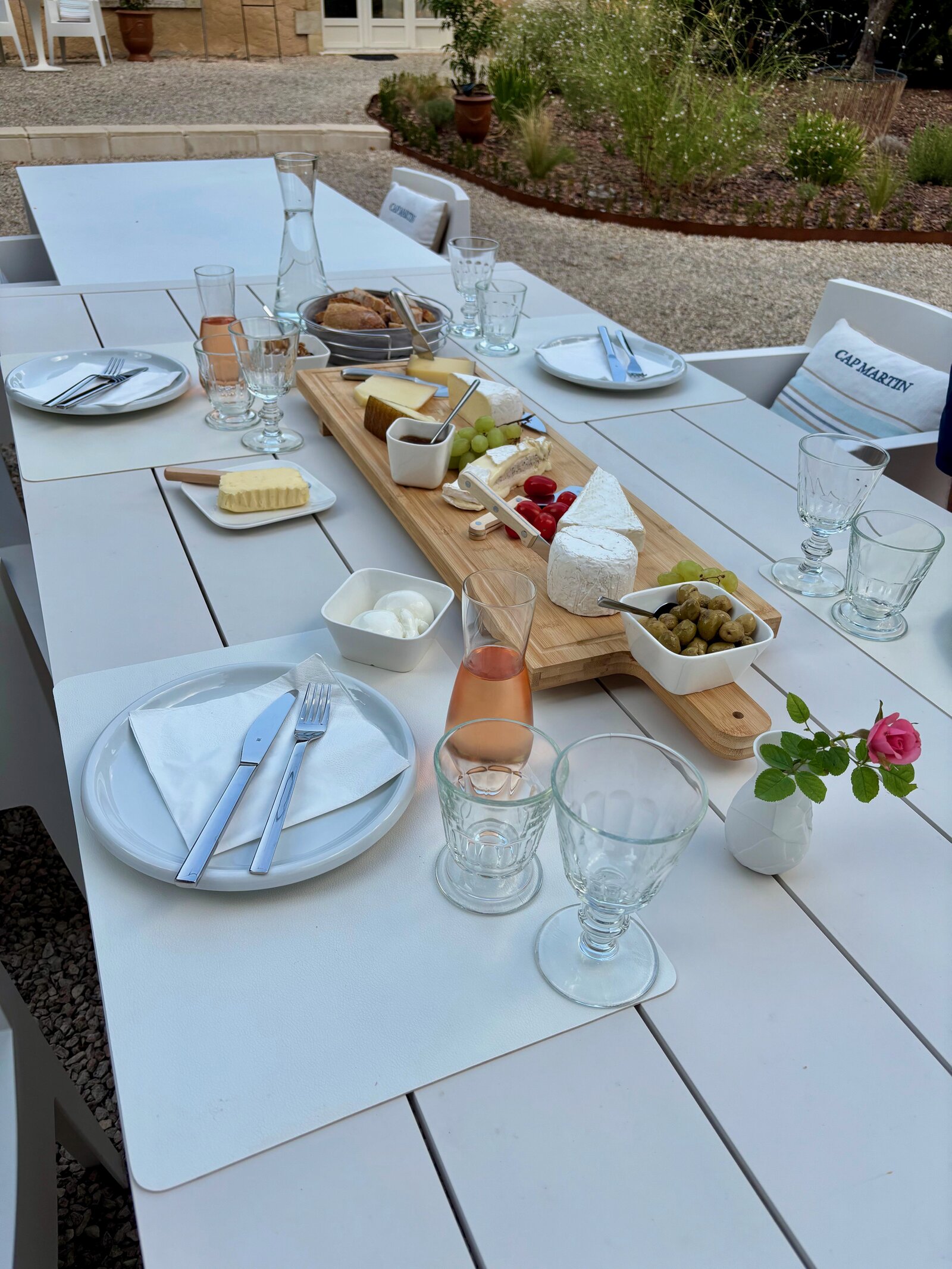 Outdoor table laid with plates, glasses of rosé, a cheese board, olives and bread, with the gravel courtyard behind.