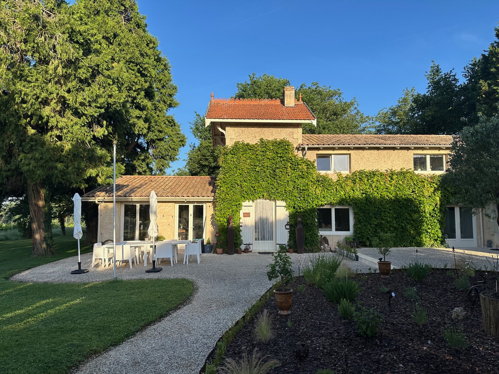 Ivy-covered stone house with a tiled roof, a gravel path and a terrace with white tables and parasols beside a lawn.