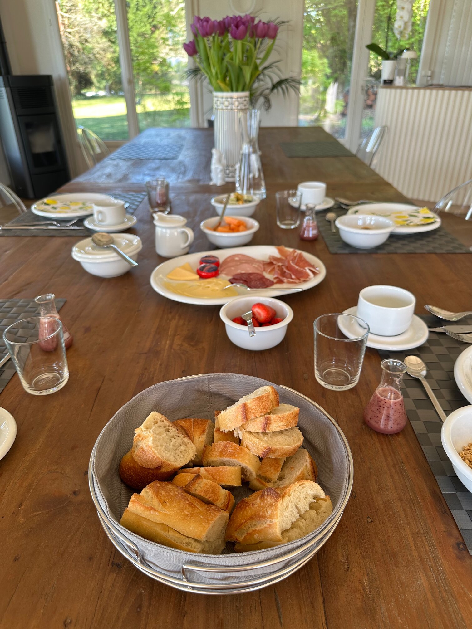 Long breakfast table with a bread basket, cured meats, fruit, jams and white cups, with a vase of tulips by the glazed wall.