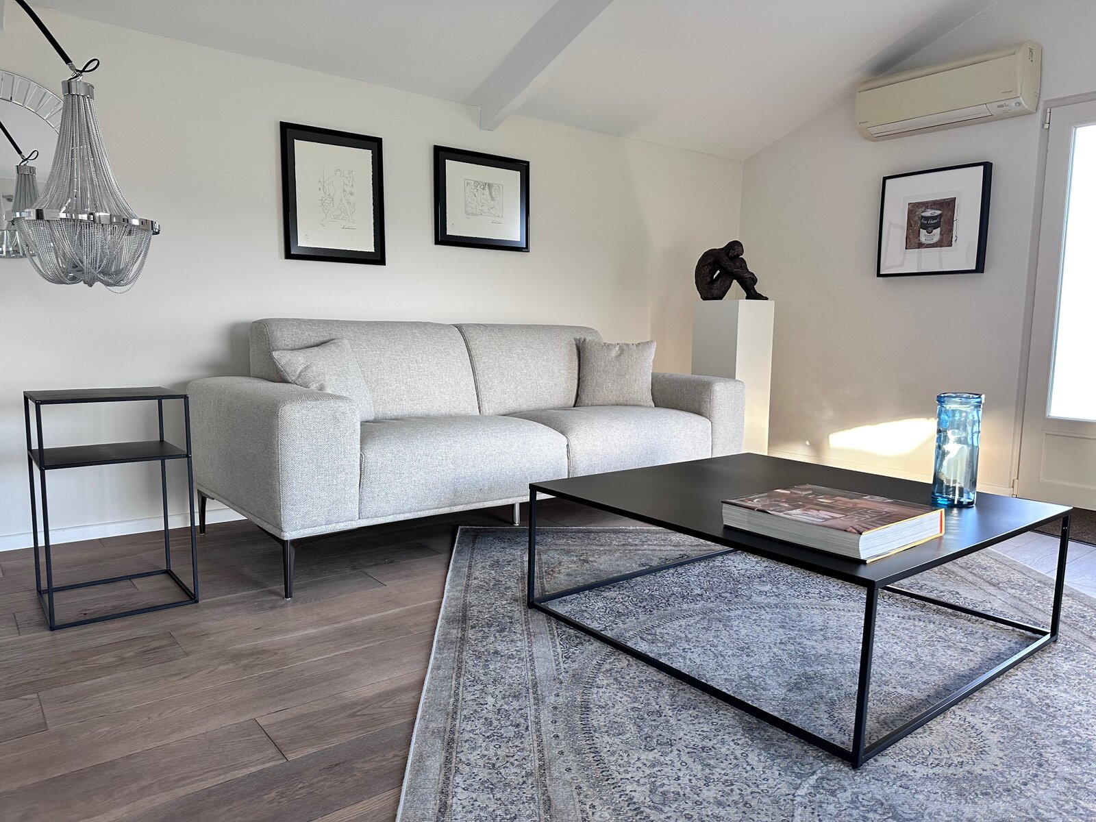 Lounge under a sloping ceiling with a light grey sofa, a black metal coffee table on a rug, framed drawings and a small sculpture on a plinth.