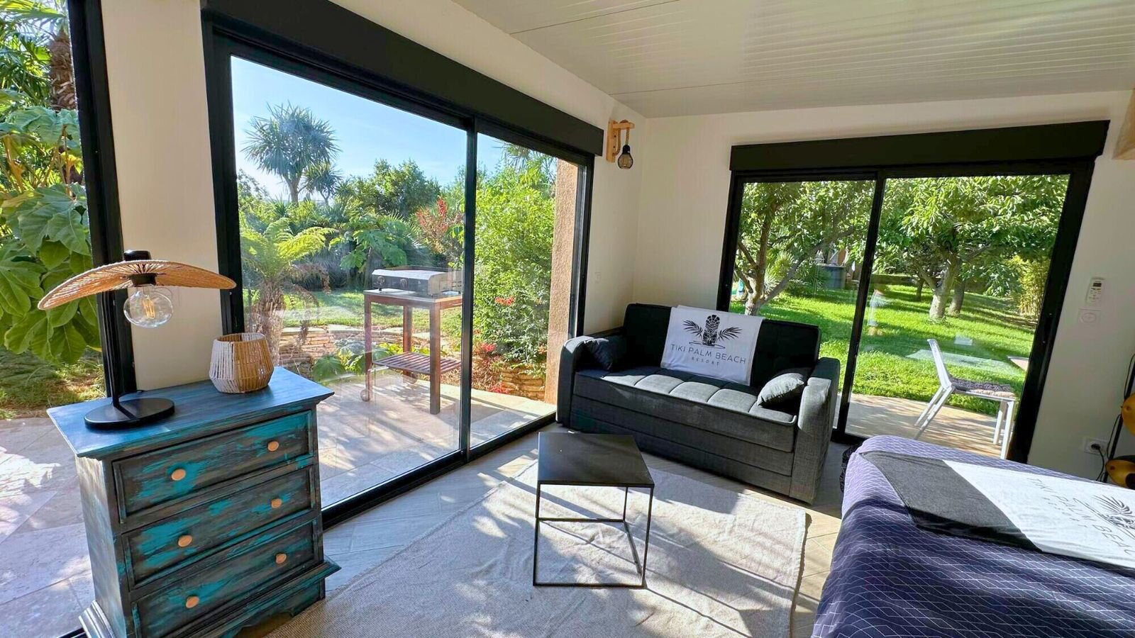 Garden room with two glass walls onto the lawn, a dark sofa with a cushion bearing the logo, a painted chest of drawers and a bed.