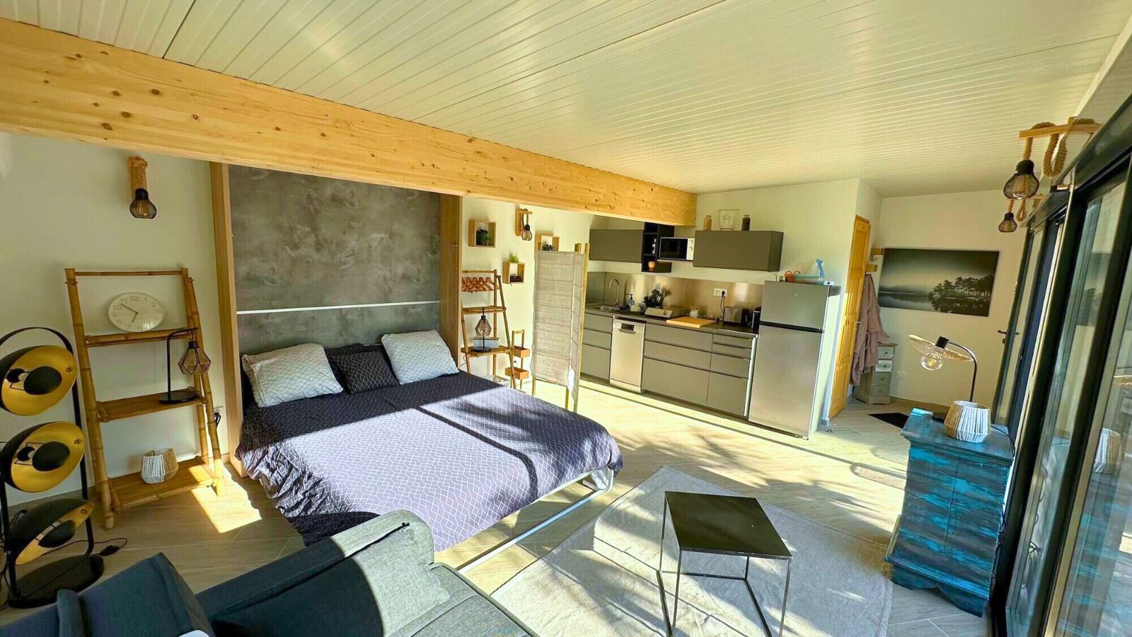 Studio with a double bed under a wooden beam, a kitchenette with a fridge along the back wall and a glass wall on the right.