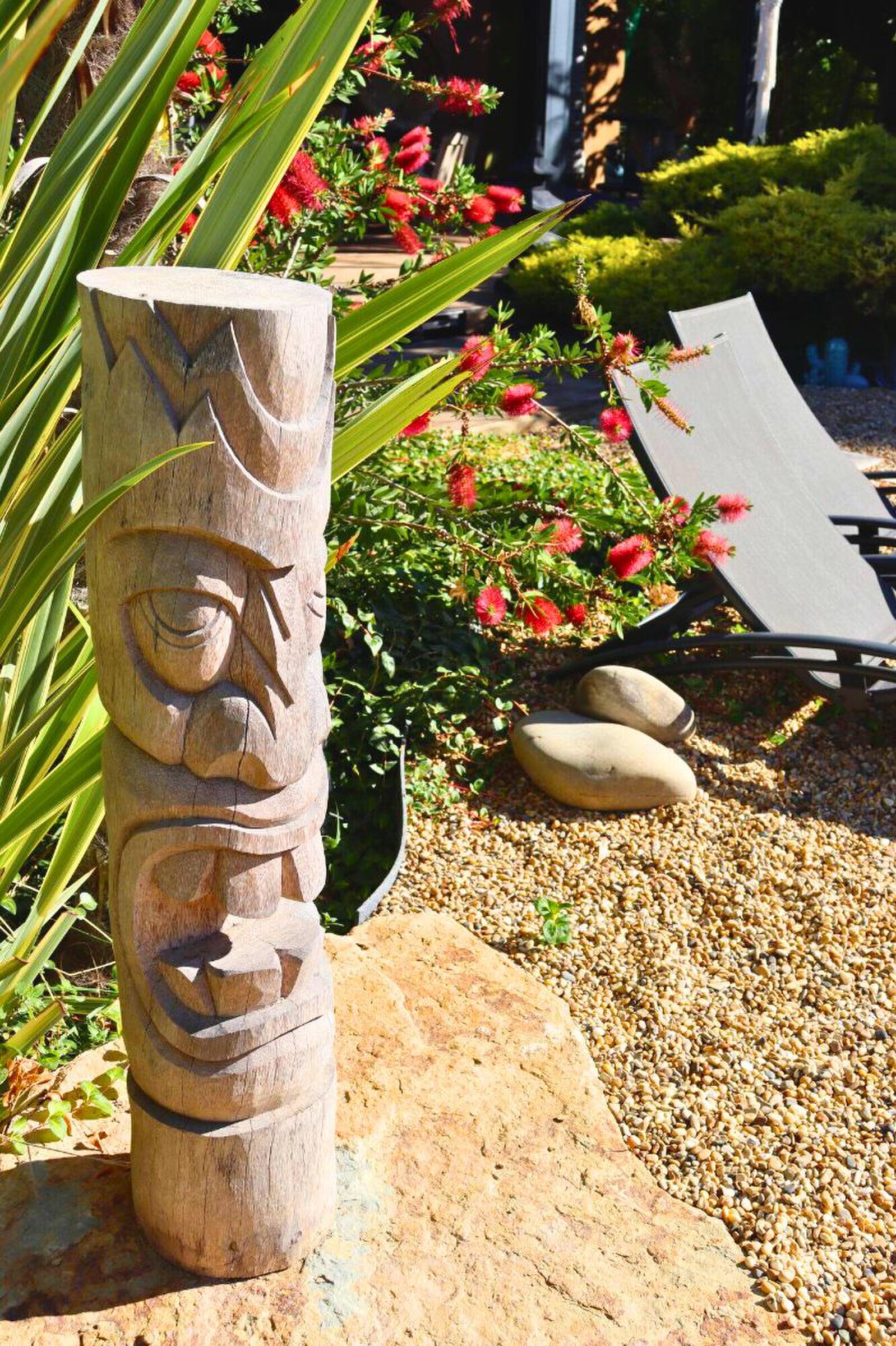 A carved wooden tiki post on a rock, with red bottlebrush flowers and a sun lounger on the gravel behind.