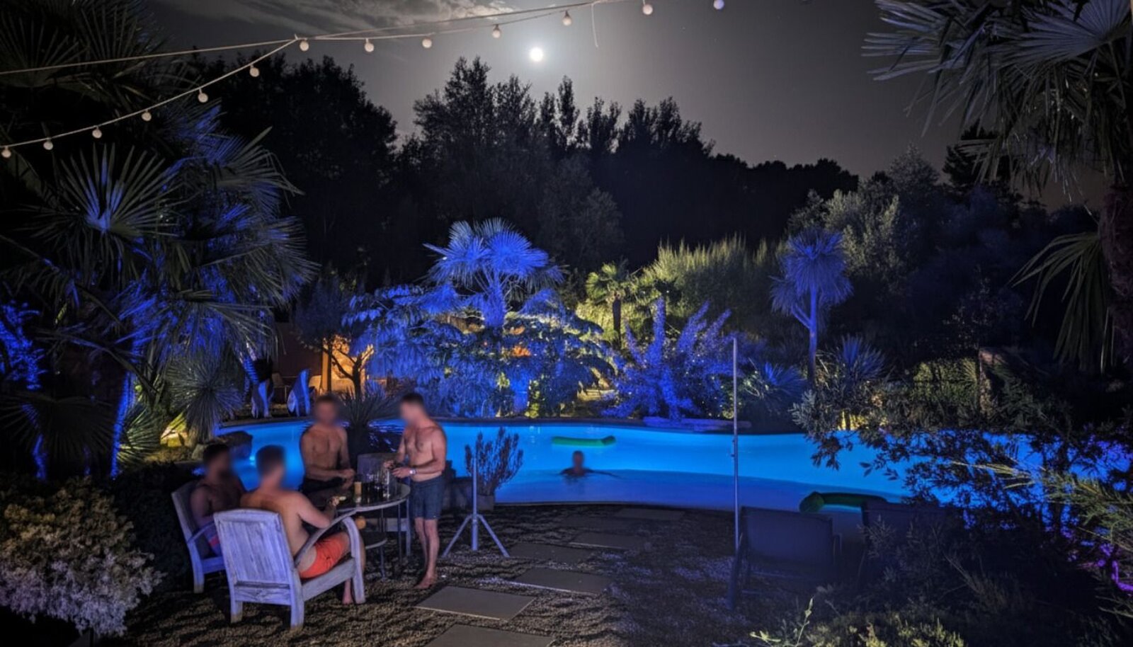 The lit pool at night under a full moon and string lights, with a few people sitting at a table by the water.