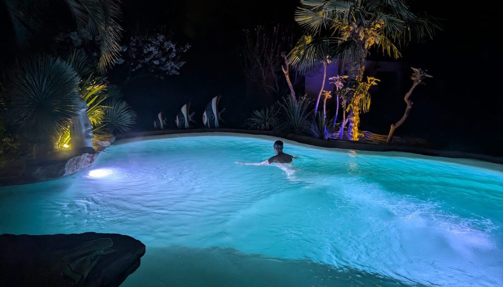 The pool lit turquoise at night, with one swimmer and lit palms and fish figures along the edge.