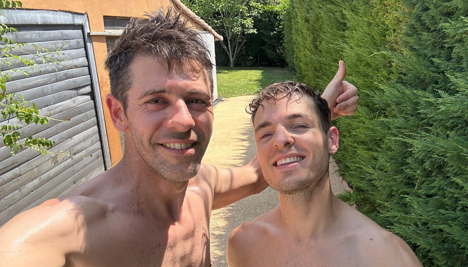 Two shirtless men smiling for a selfie on a garden path beside a hedge.