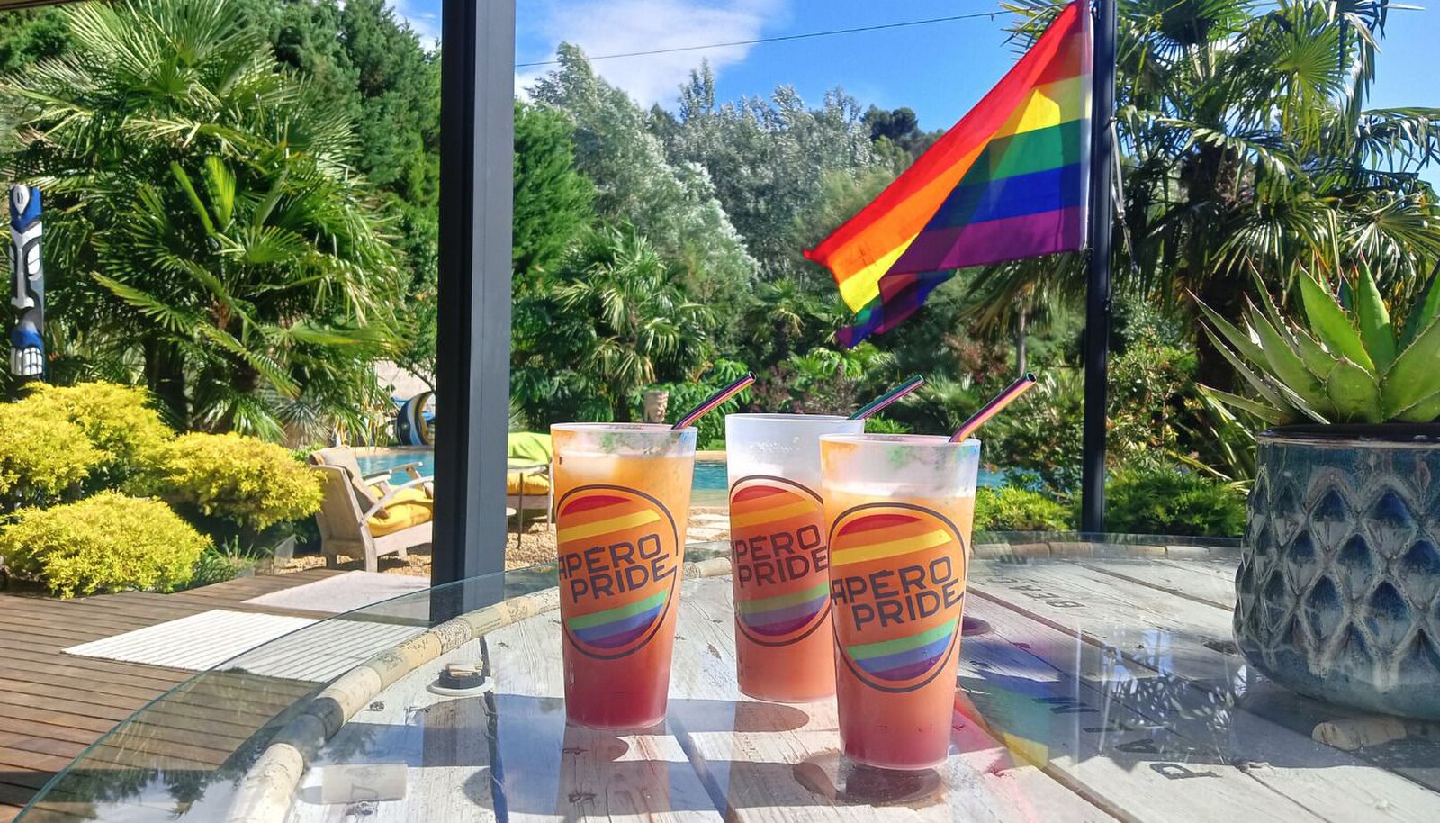Three cocktails in rainbow-printed cups on a glass table, with a rainbow flag, palms and the pool behind.
