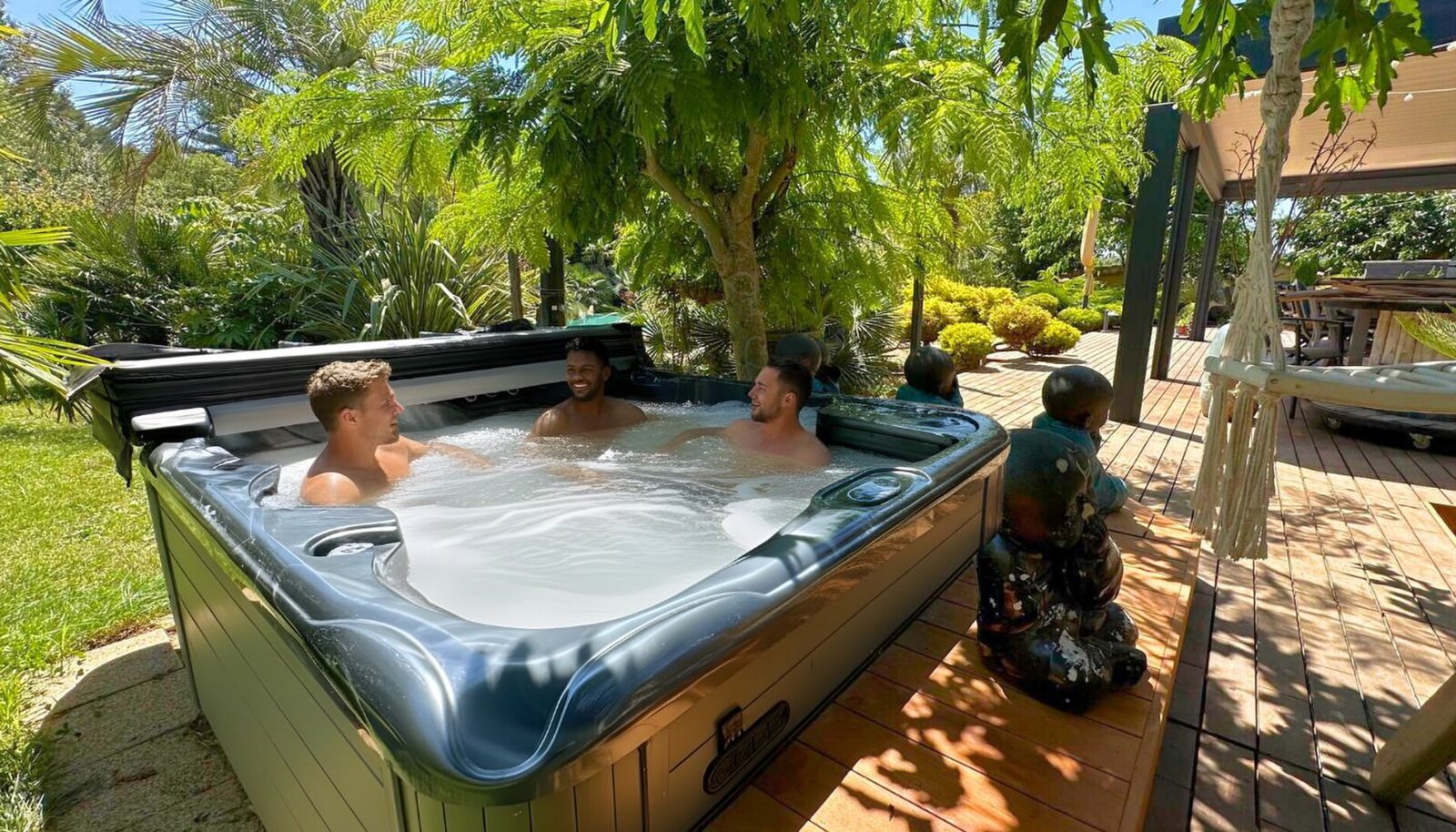 Three men in a hot tub on a wooden deck, surrounded by trees and Buddha figures.
