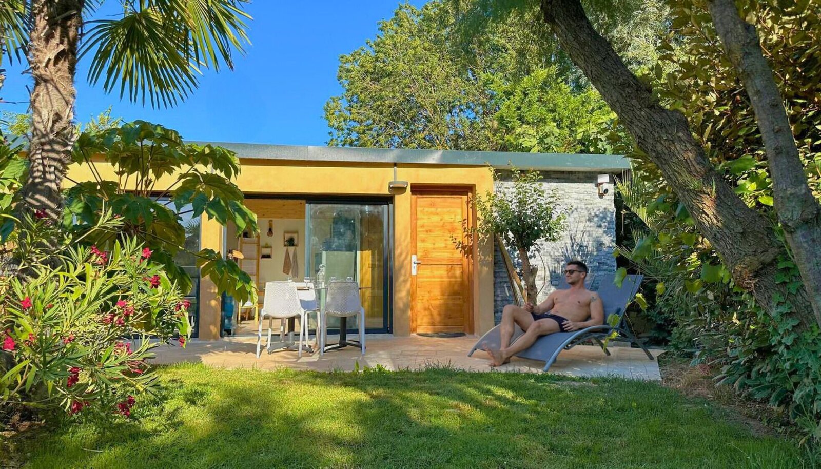 A small yellow garden house with a glass door and a table outside, and a man lying on a lounger on the lawn in front.