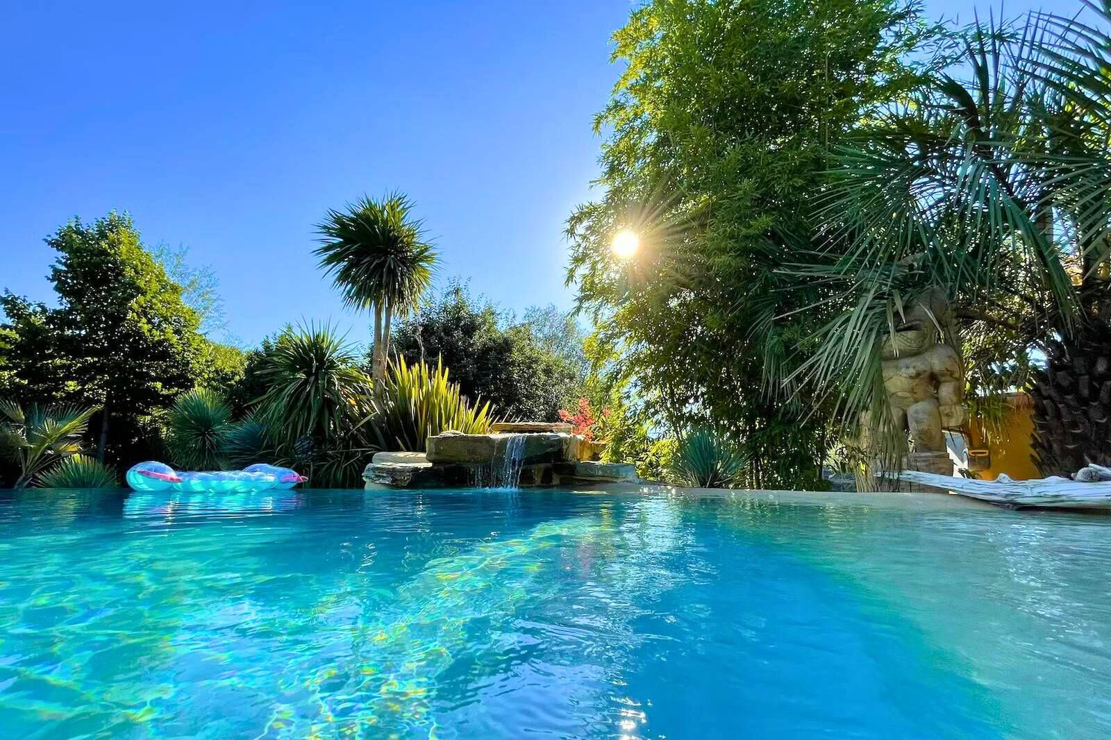 The pool at water level, with a small stone waterfall, palm trees and a carved statue in the sunshine.
