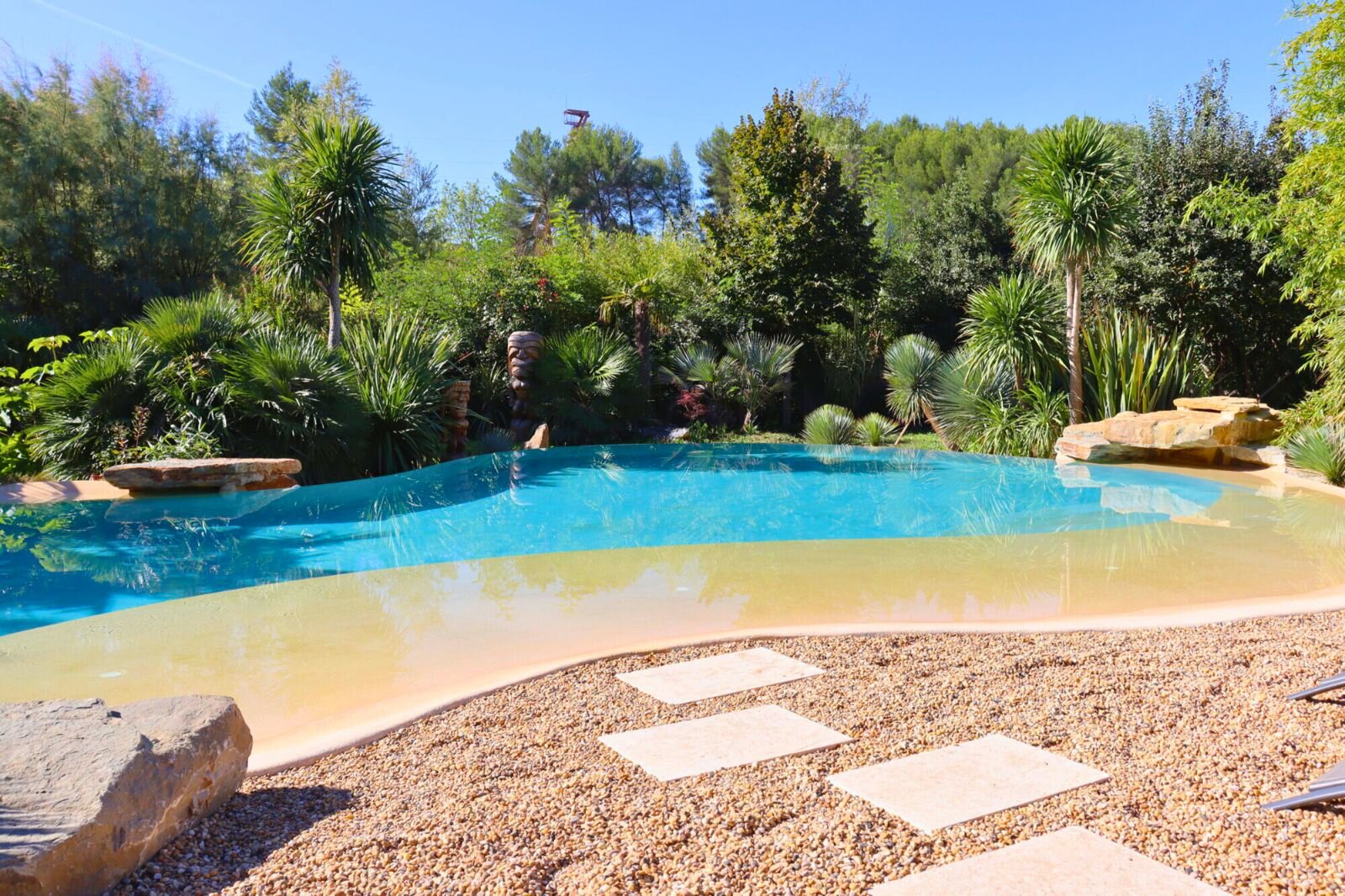 A free-form pool with a sandy shallow edge, stepping stones across the gravel and palms and shrubs around it.