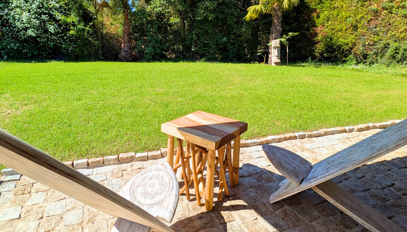 A small wooden stool between two wooden deck chairs on a cobbled terrace, facing a lawn and a garden hedge.