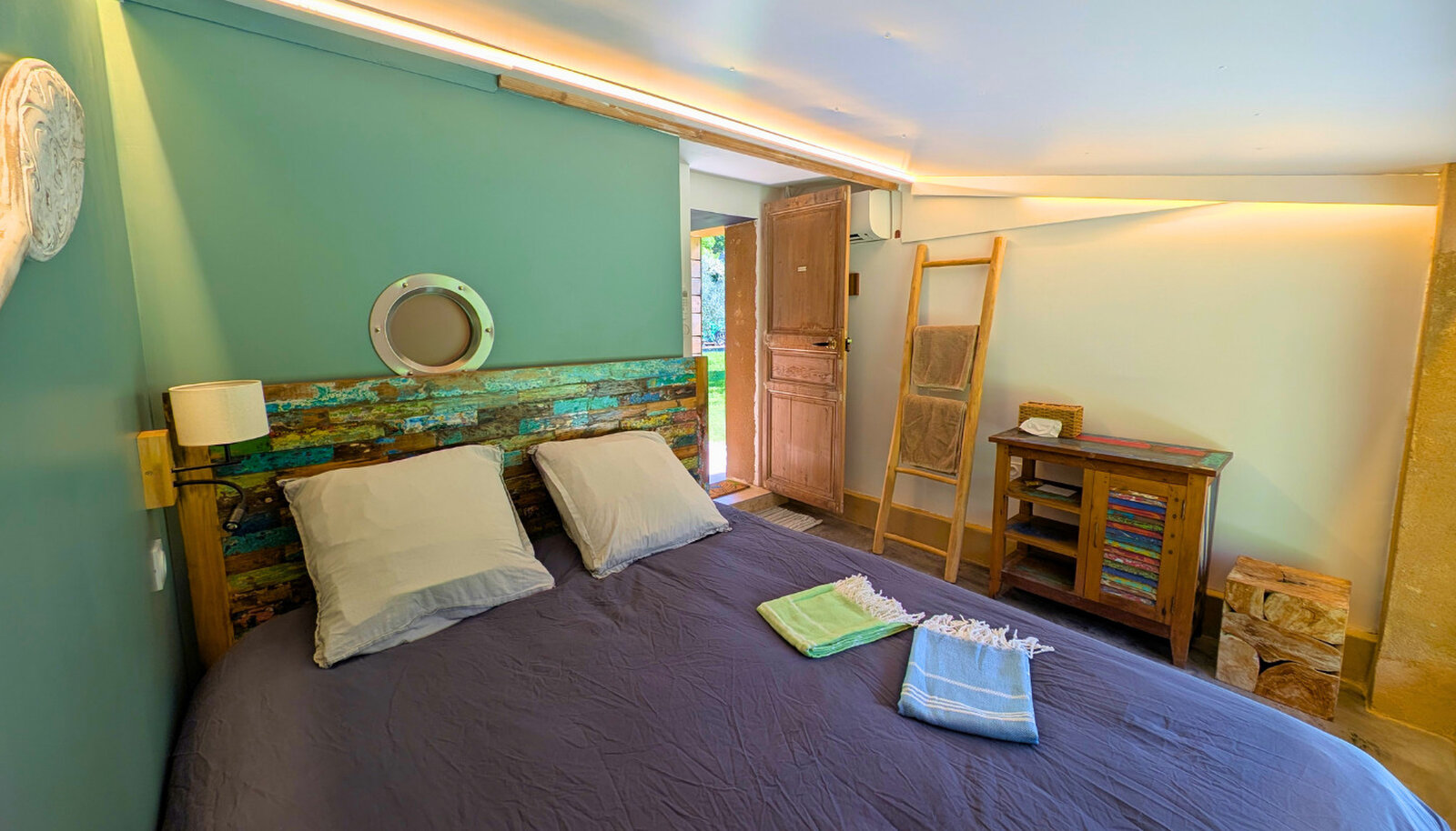 Bedroom with a reclaimed painted wood headboard, a porthole mirror, a towel ladder and folded towels on a dark bedspread.