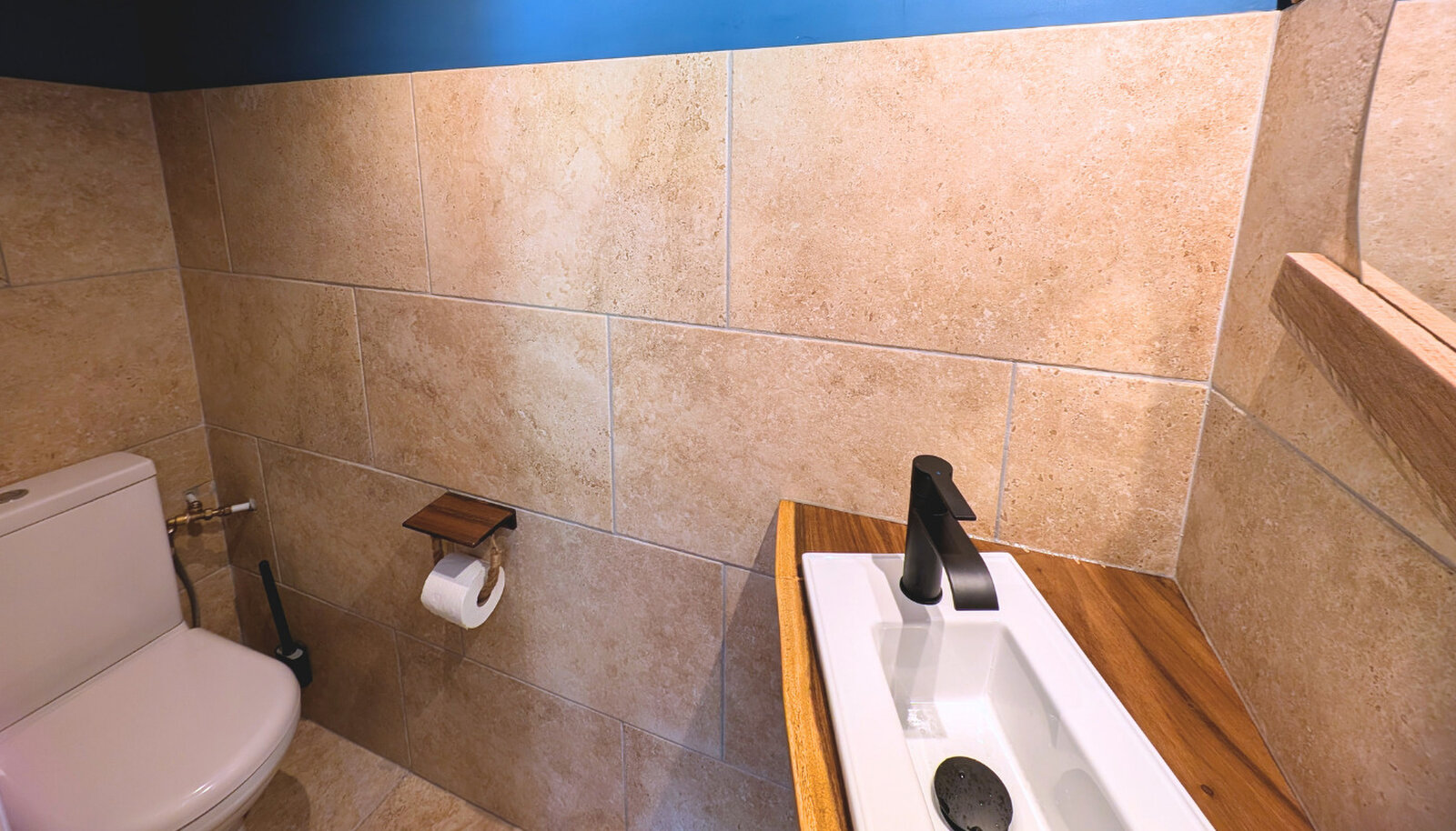 A toilet with stone-look wall tiles, a wooden shelf and a small white basin with a black tap.