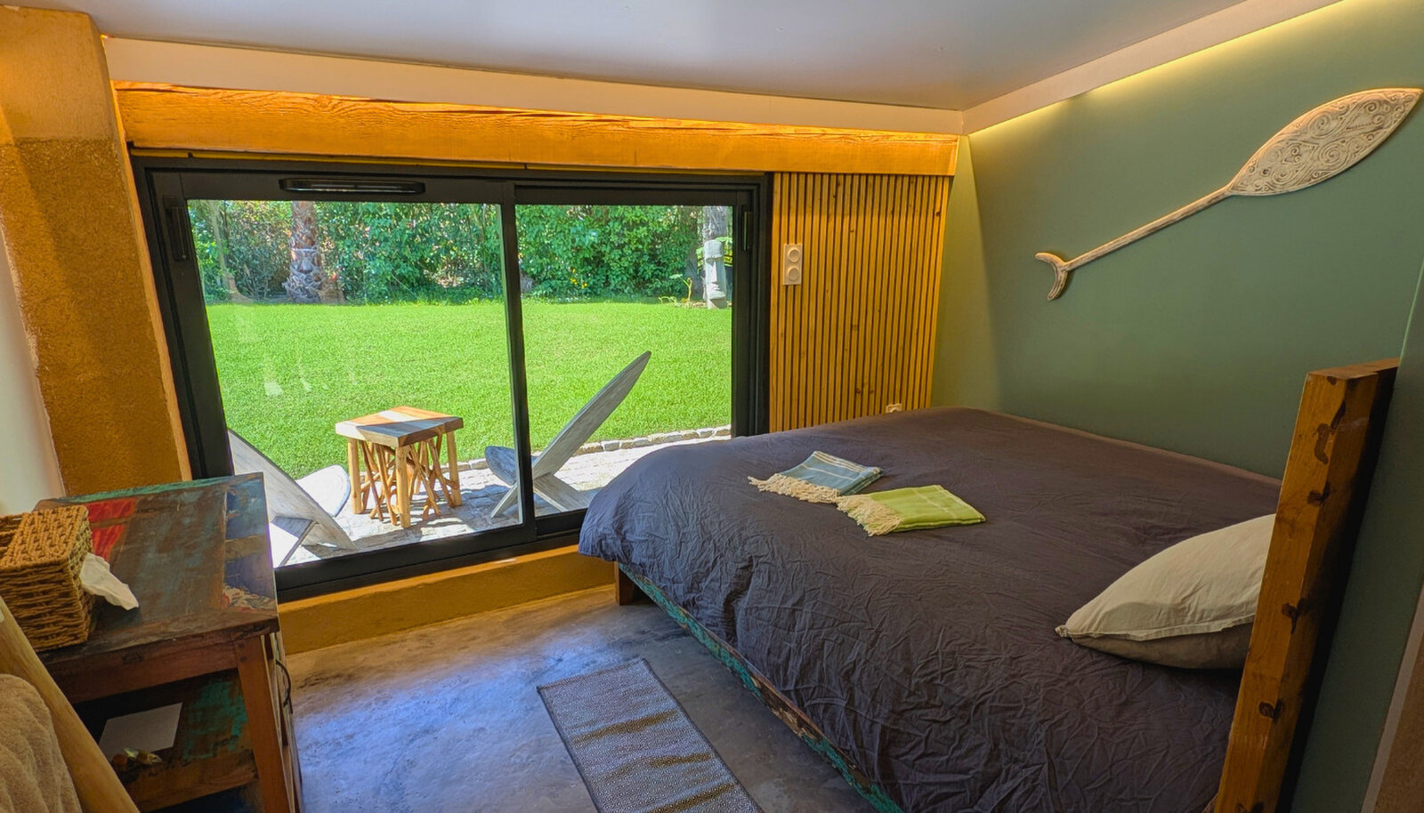 Bedroom with a glass sliding door onto the lawn and terrace, a dark bedspread with folded towels and a carved paddle on the green wall.