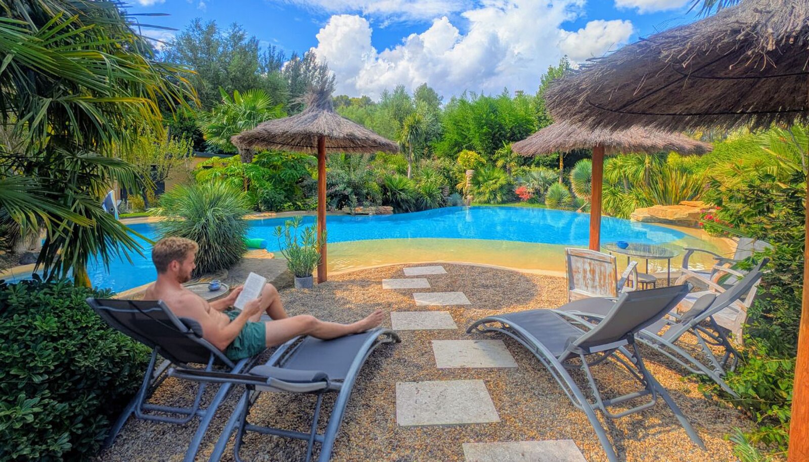 A man reading on a sun lounger by the pool, with thatched parasols and more loungers on the gravel.