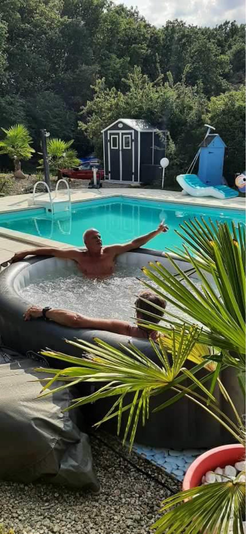 Two men relaxing in an inflatable hot tub beside the swimming pool, with a garden shed and trees behind.