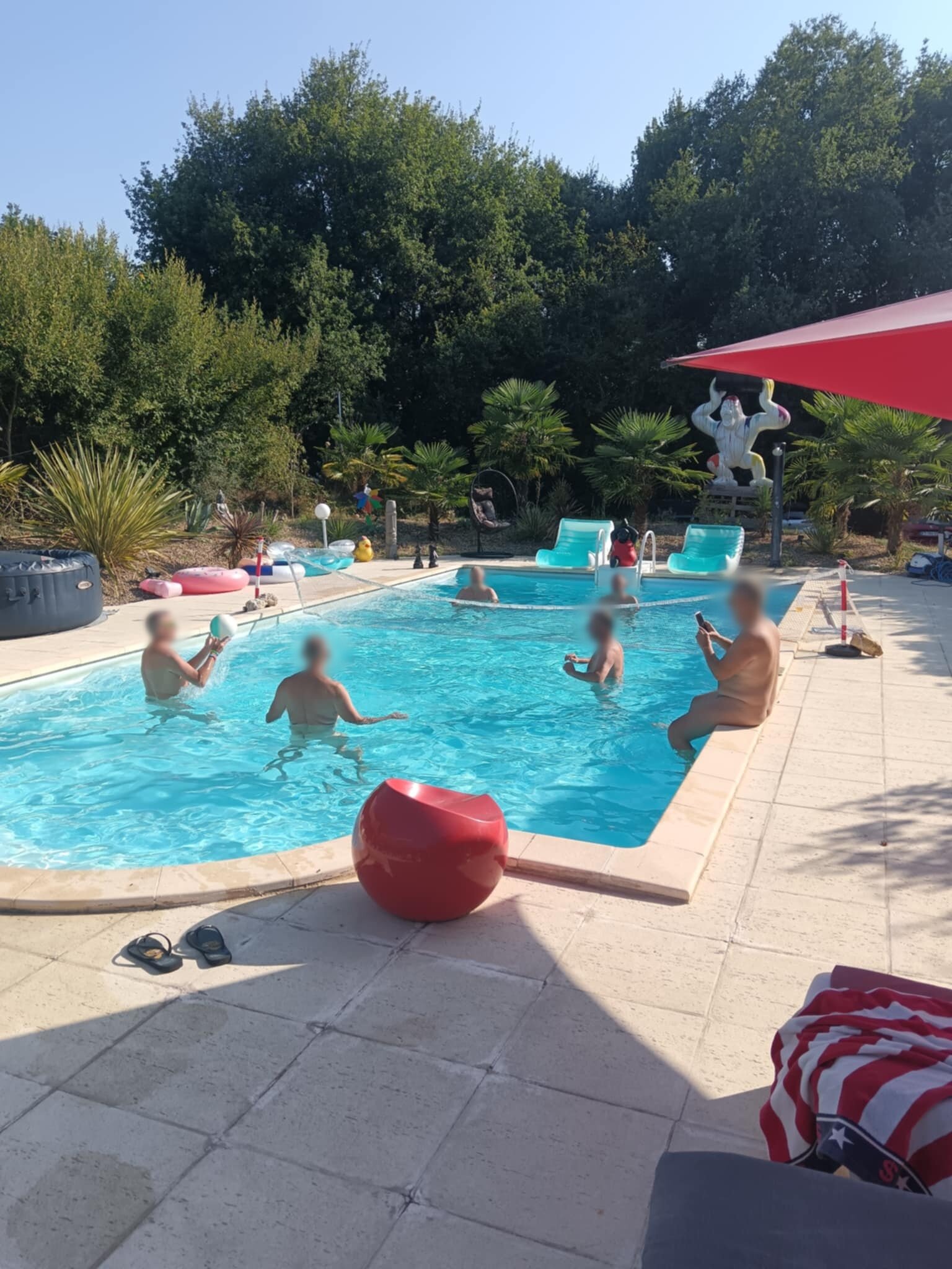 Guests playing in the swimming pool, with a red stool on the paving and a large statue among the palms at the far end.