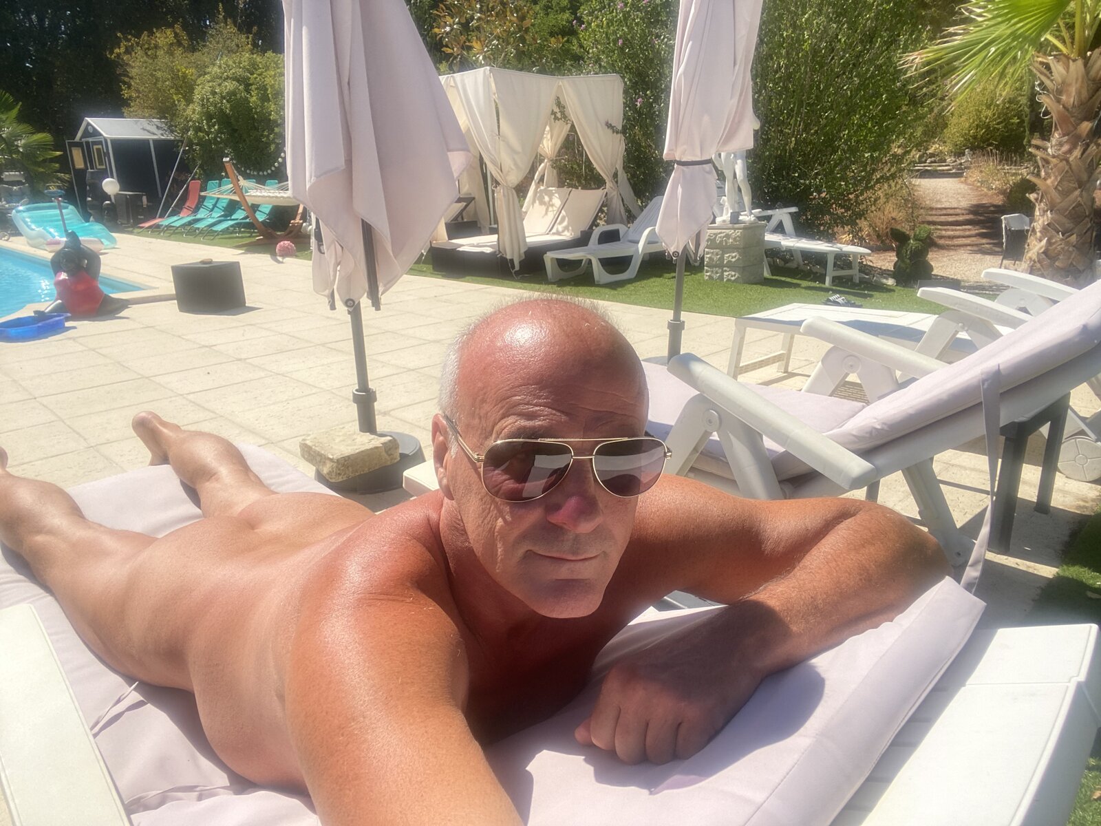 A naked man lying on a cushioned sun lounger by the pool, with parasols and a canopy bed behind.
