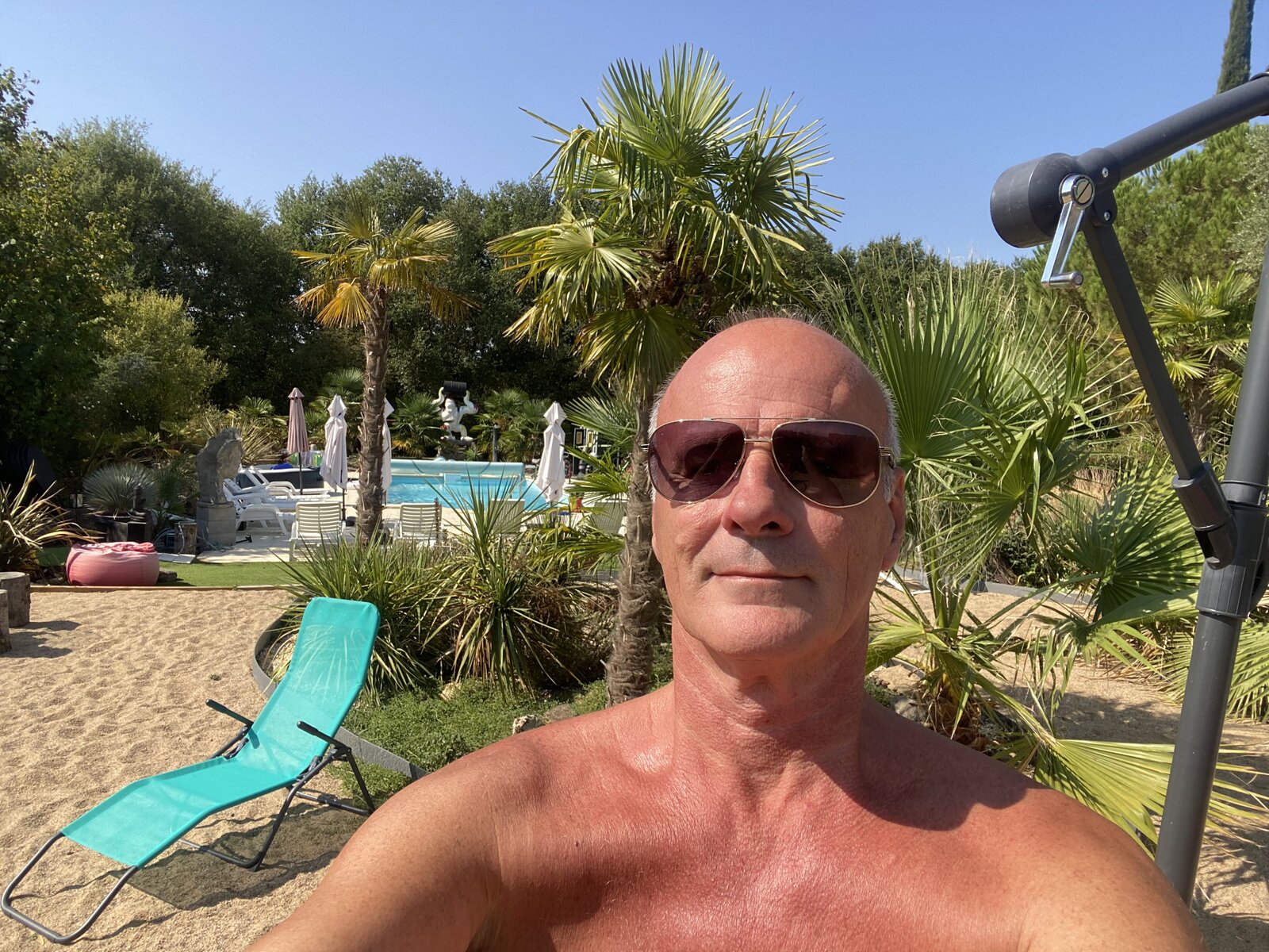 A shirtless man in sunglasses taking a selfie, with palms, loungers and the pool in the garden behind.