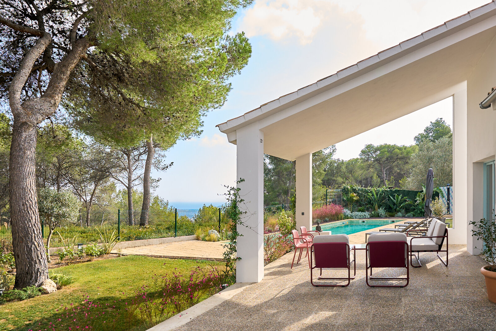 A covered terrace with red armchairs looking onto the pool and the garden, with a sea view between the pines.