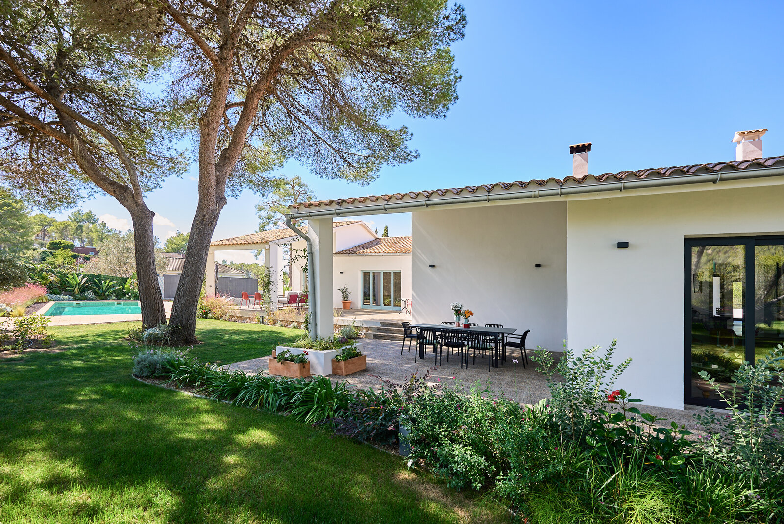 The garden with a lawn under tall pines, a dining table on the terrace and the pool in the distance.